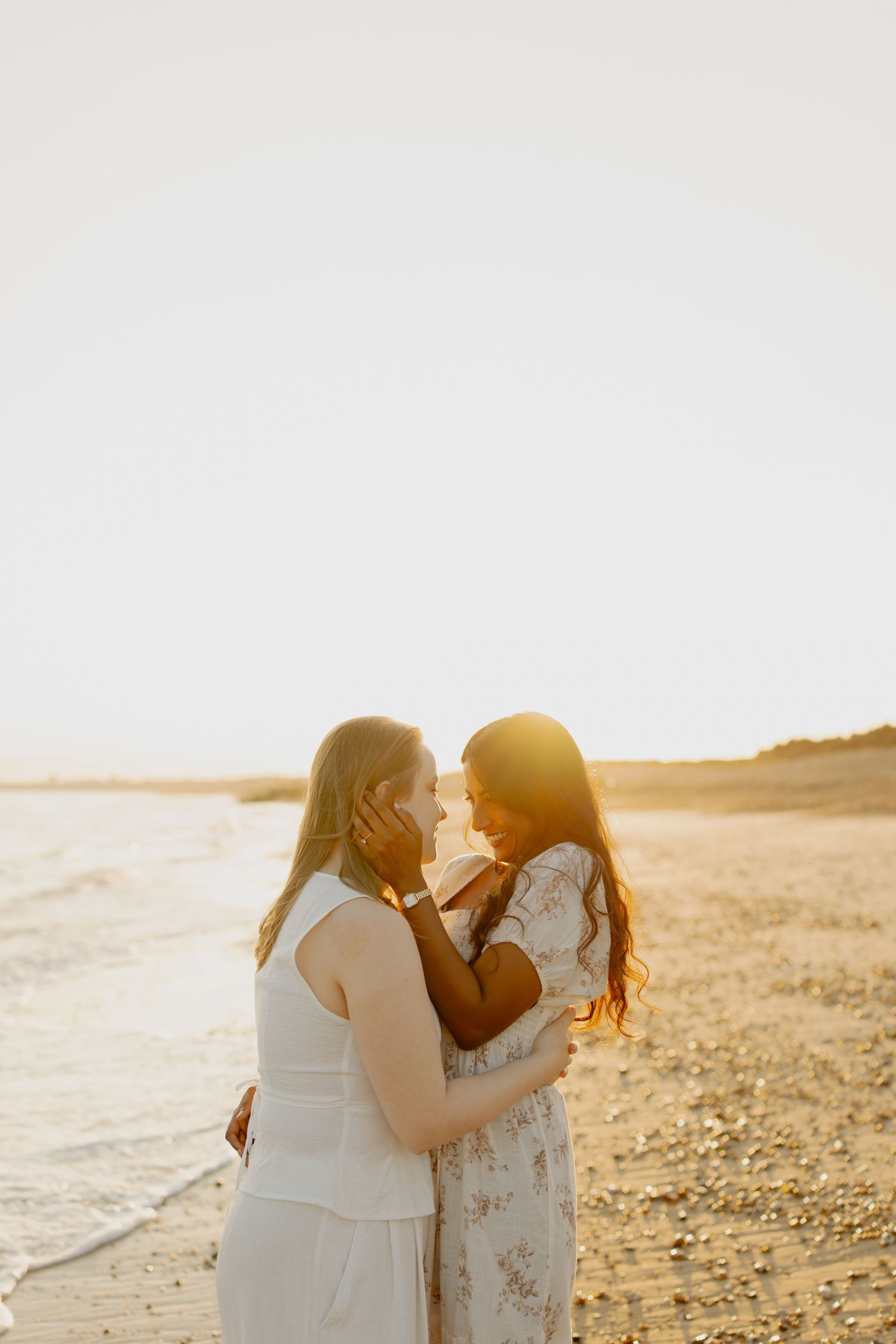 Two women in white dresses embrace on a sandy beach at golden sunset, sharing a romantic moment by the ocean.