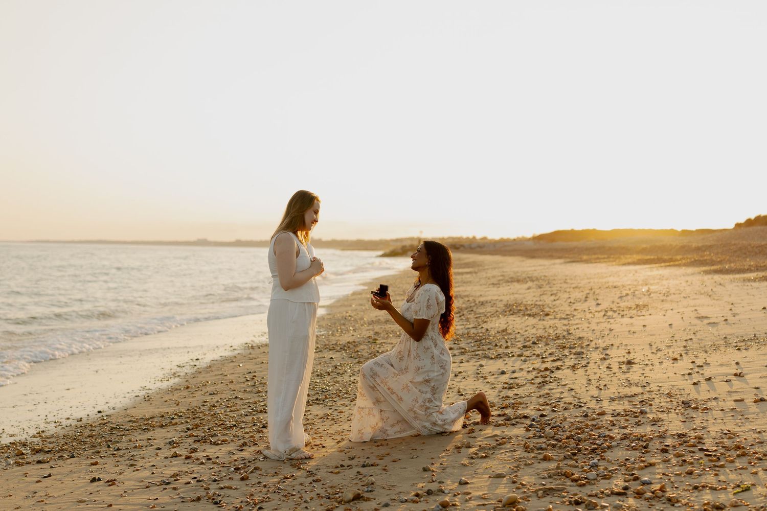 Same-sex beach proposal at sunset, woman kneeling with ring box before partner on sandy shore.
