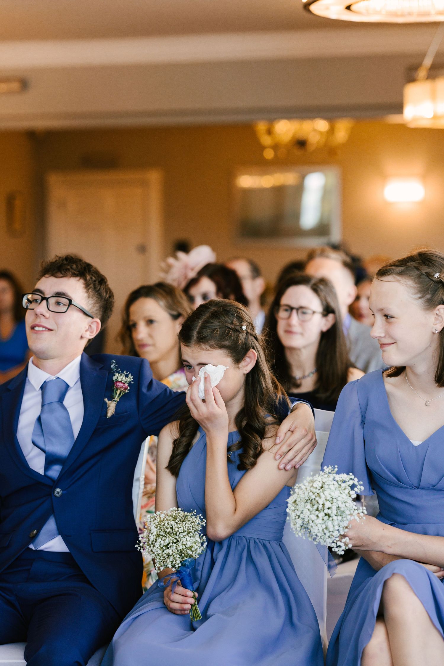 Wedding guests in blue attire react emotionally during ceremony while holding baby's breath bouquets.