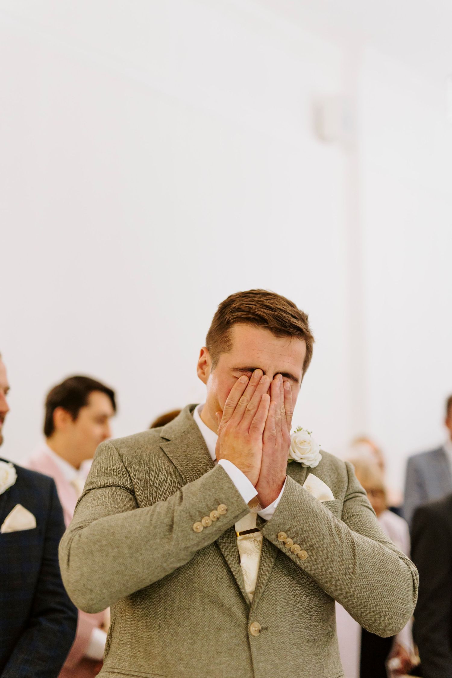 Emotional groom in tan suit covers face during wedding ceremony.