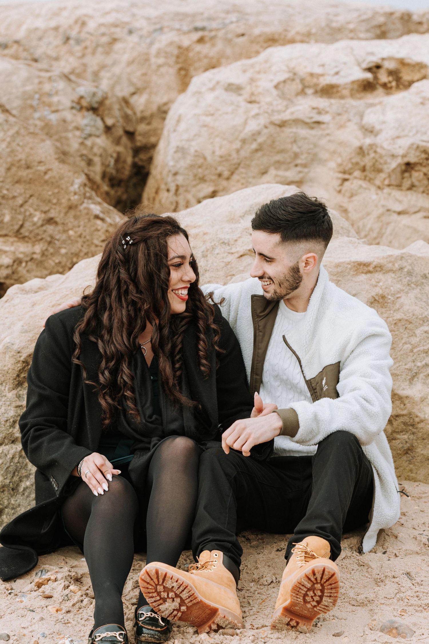 A couple sits together on coastal rocks, wearing winter clothing and sharing a joyful moment.