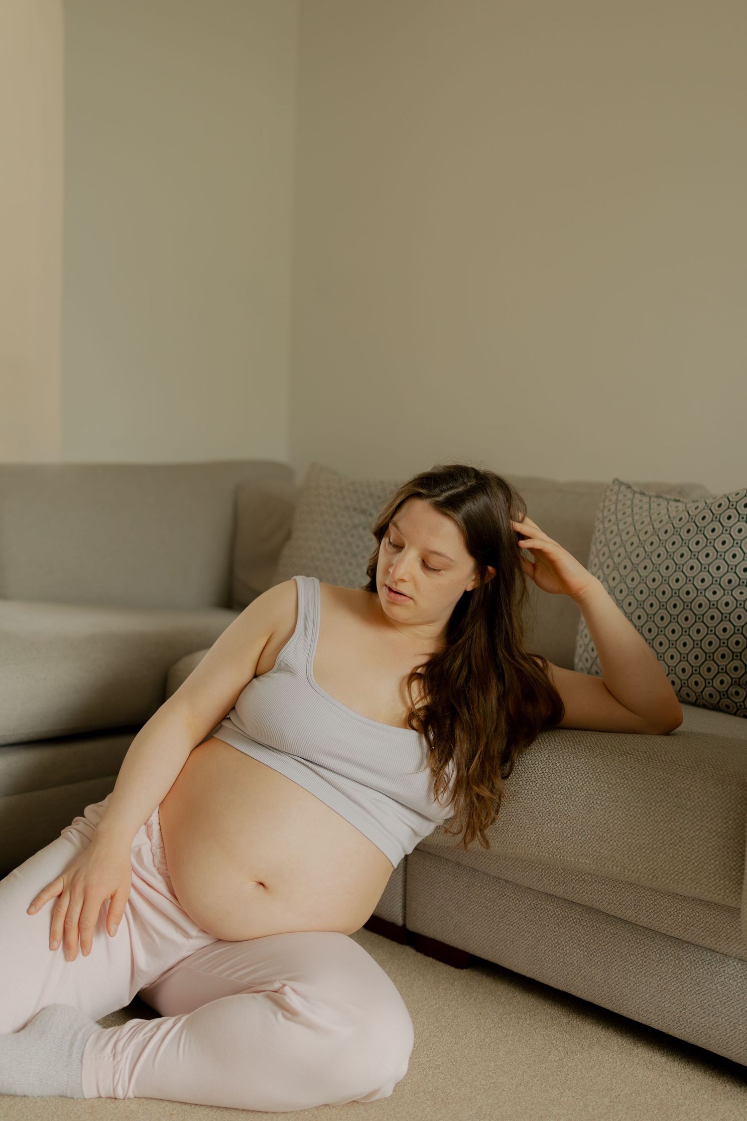 Pregnant woman sitting on floor leaning against sofa, cradling baby bump in cozy home setting.