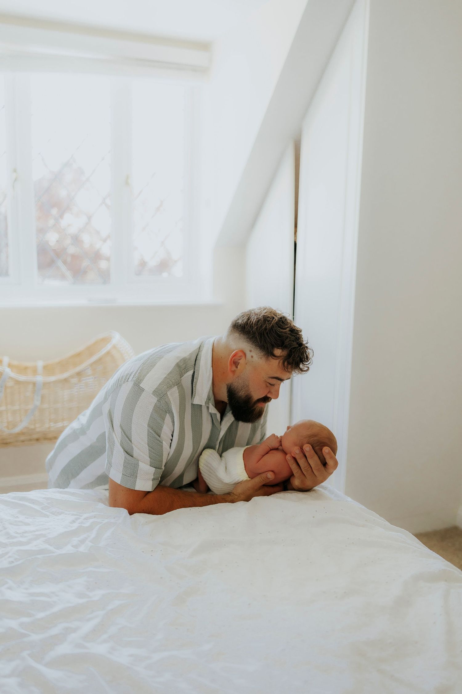 A tender moment between parent and baby captured while lying on a white bed in a sunlit room.