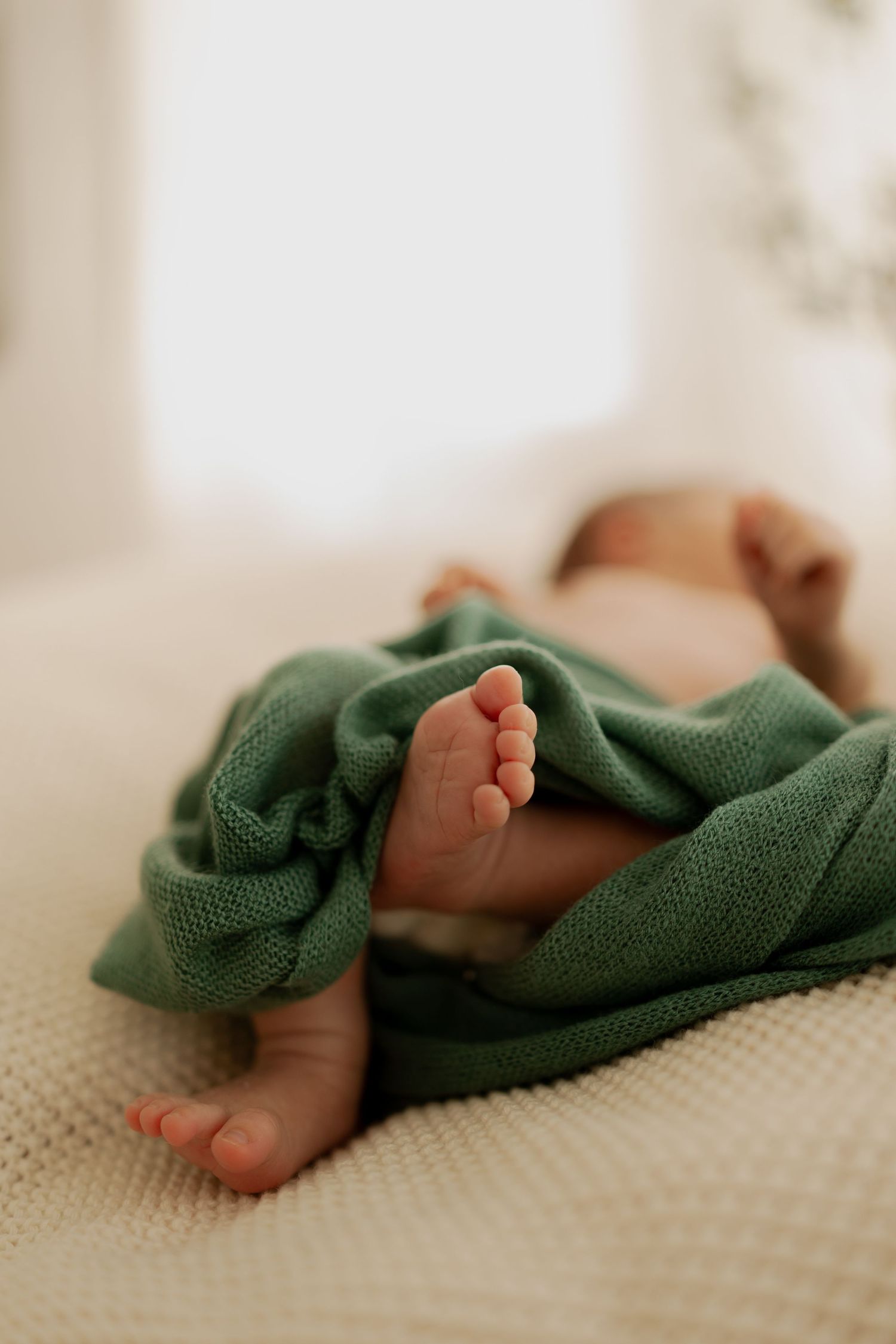 Tiny baby feet peek out from under a soft green blanket while resting on a cream-colored textured surface.