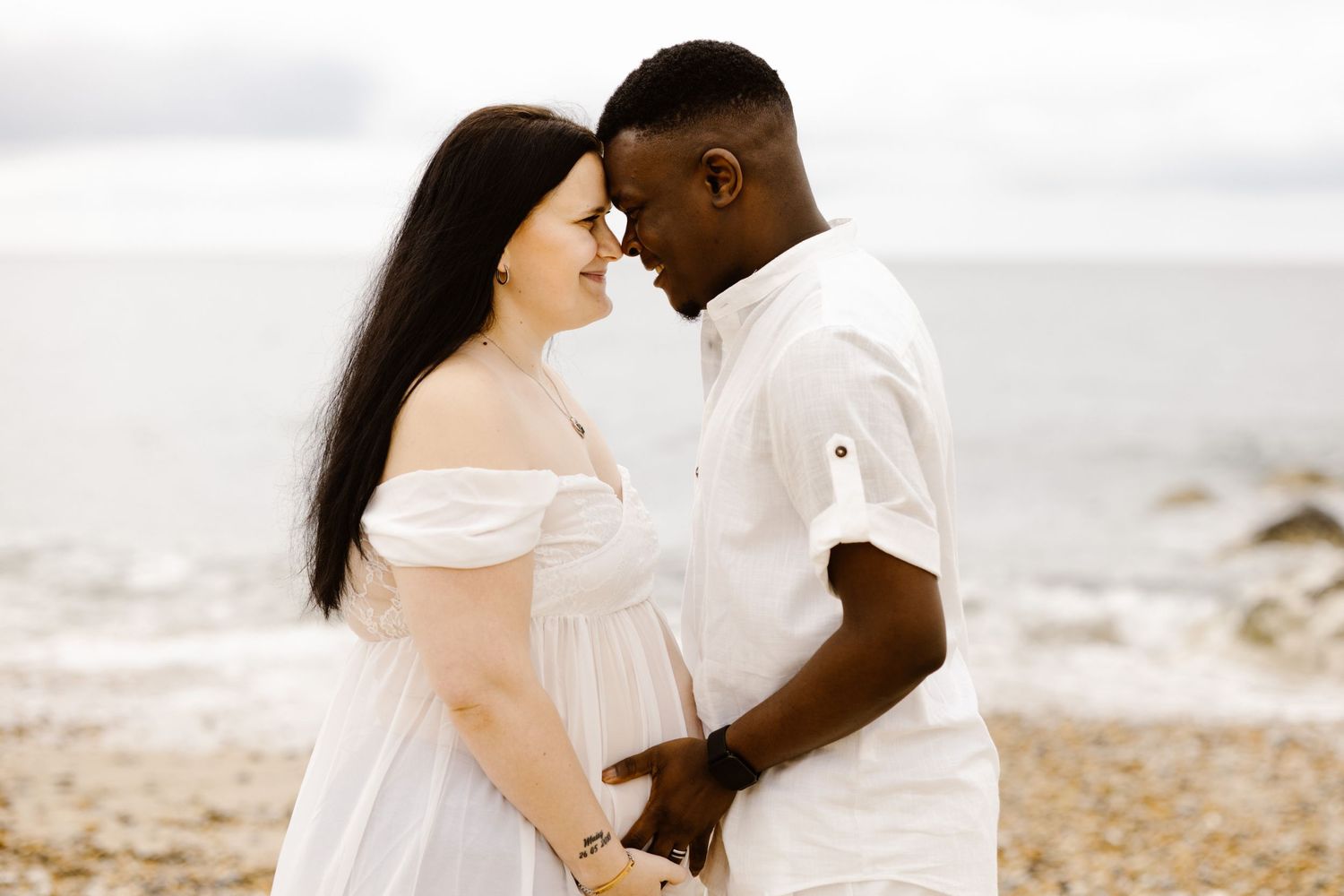 An intimate beach portrait of a couple in white outfits against an overcast sky.