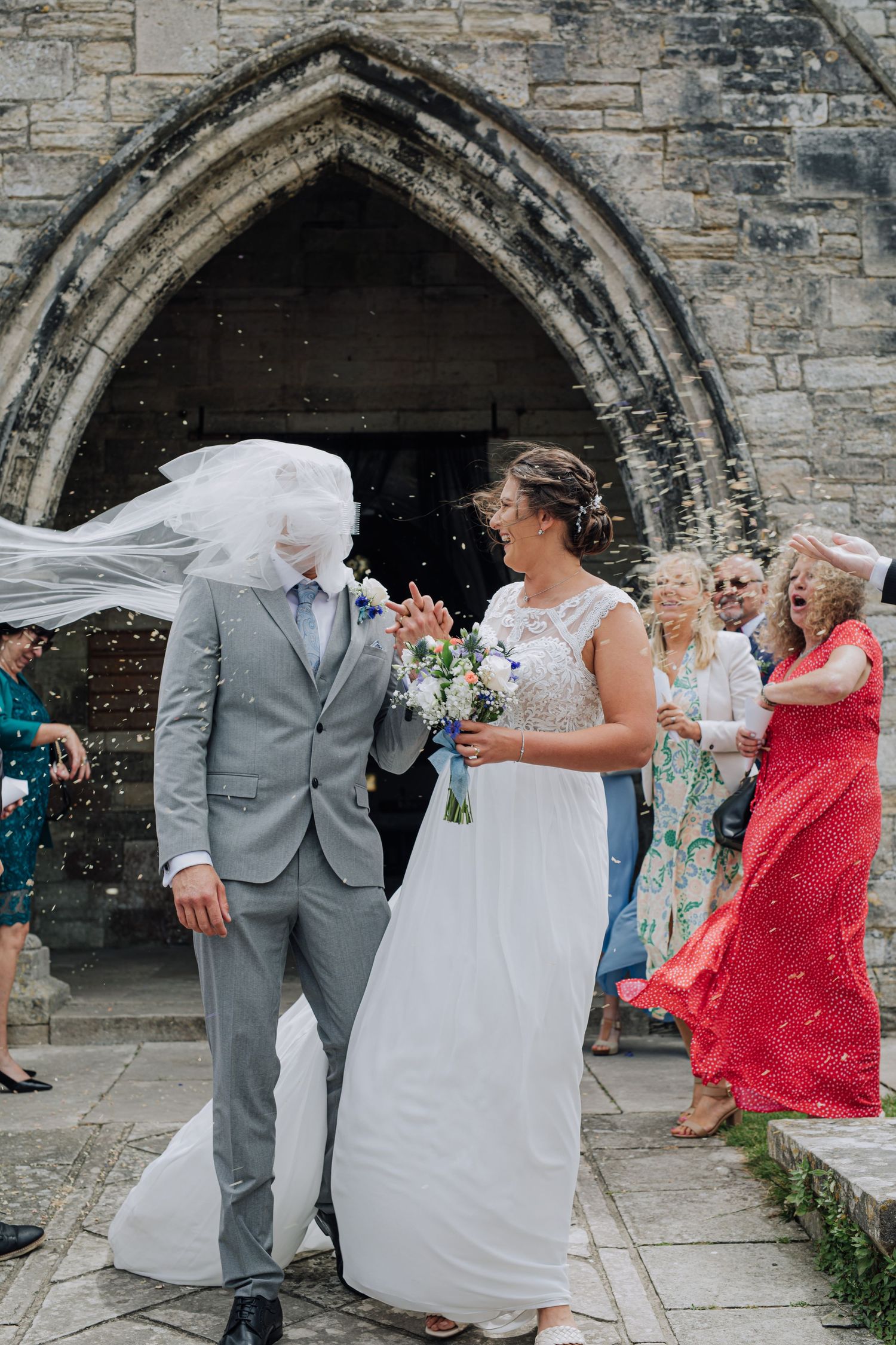 Couple celebrates outside historic stone church as wedding guests throw confetti in joyful moment.