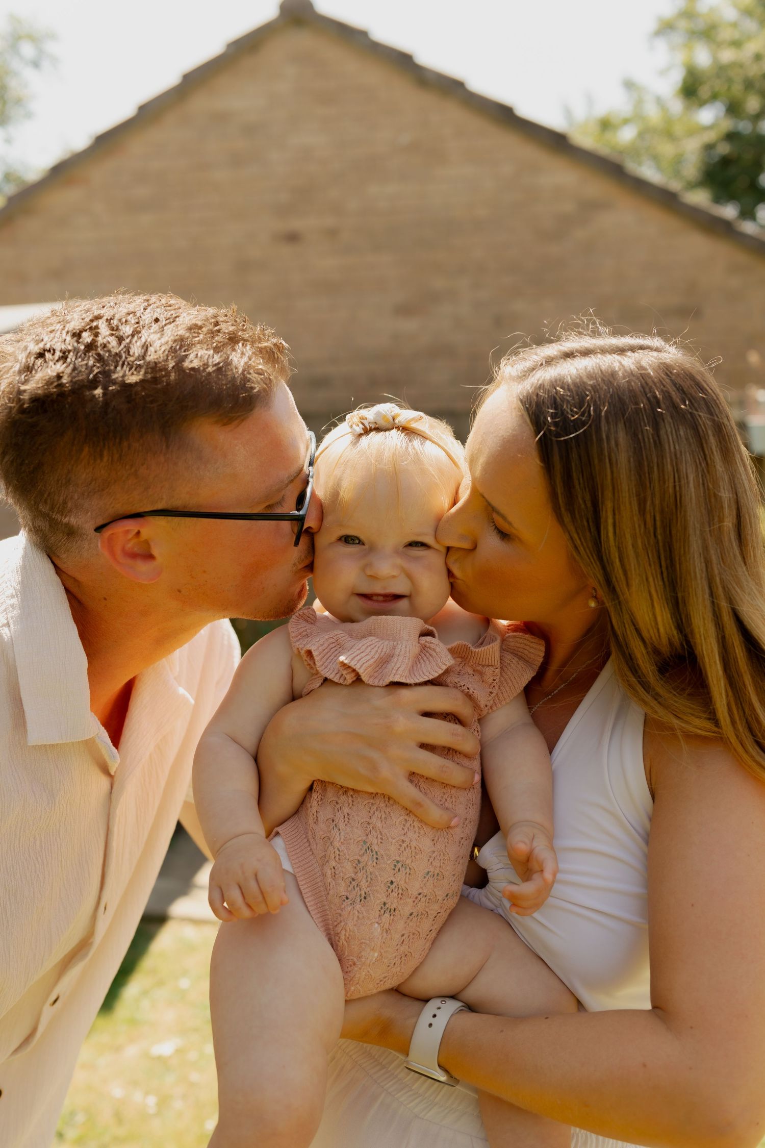 Parents kissing their smiling baby girl on the cheeks outdoors in warm sunlight near a stone building.