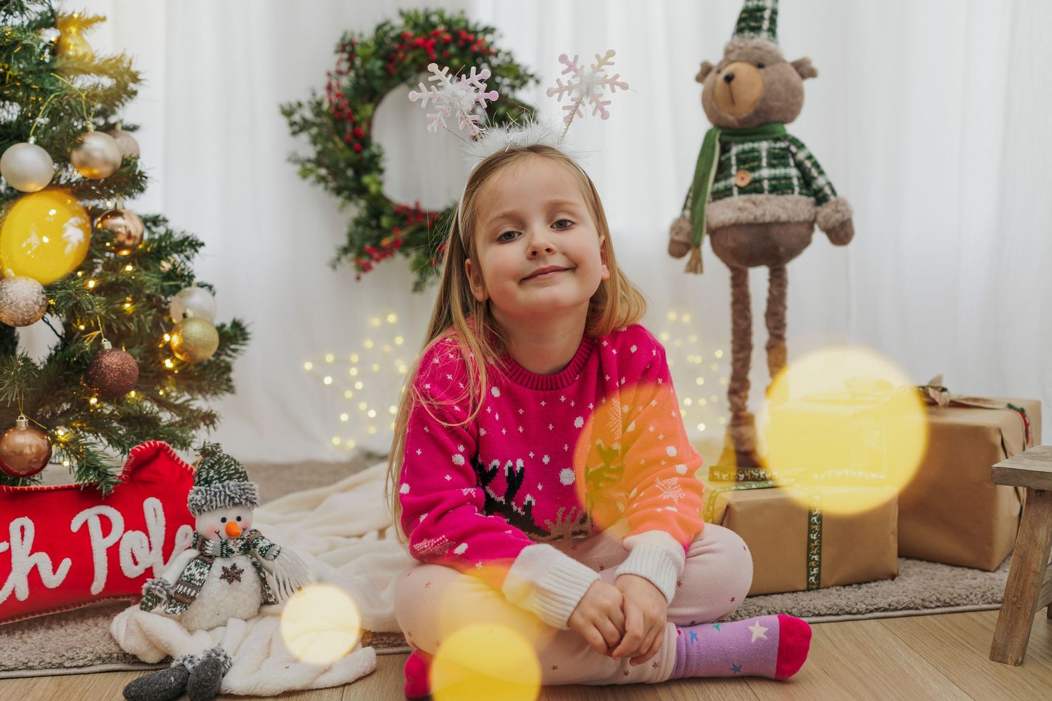 Child in pink sweater sitting among Christmas decorations with glowing string lights.