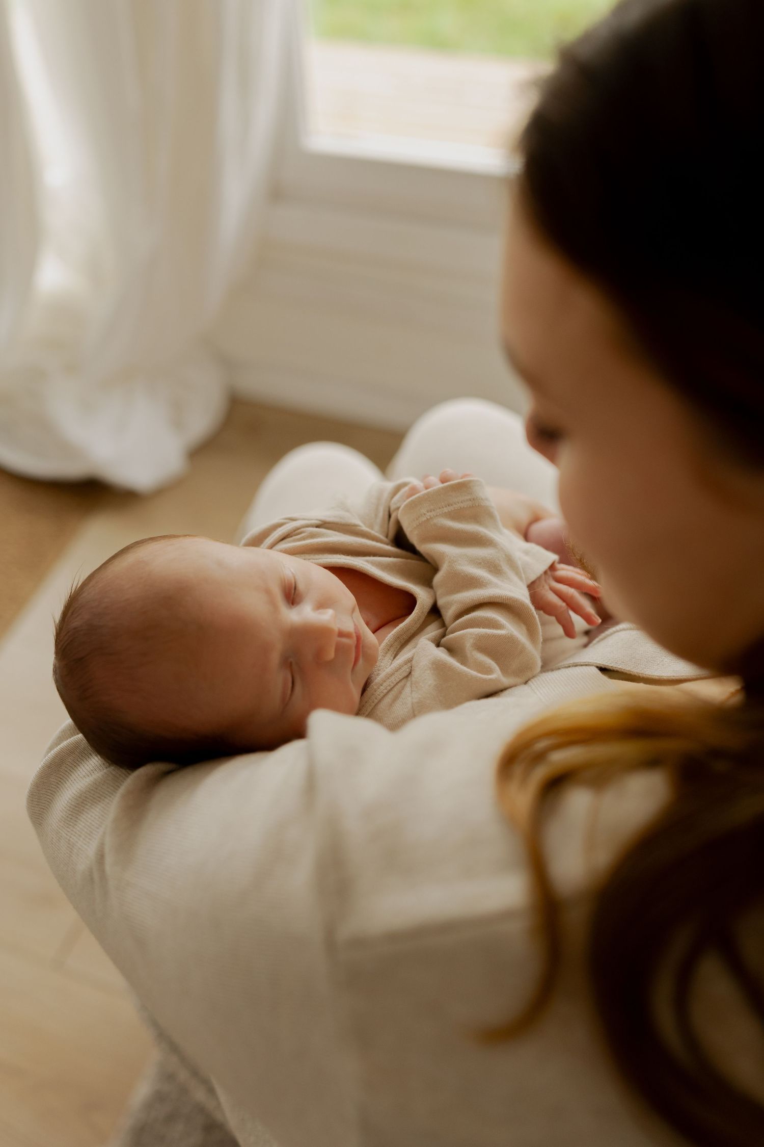 A sleeping newborn baby wearing a beige onesie rests peacefully in soft white fabric by a window.