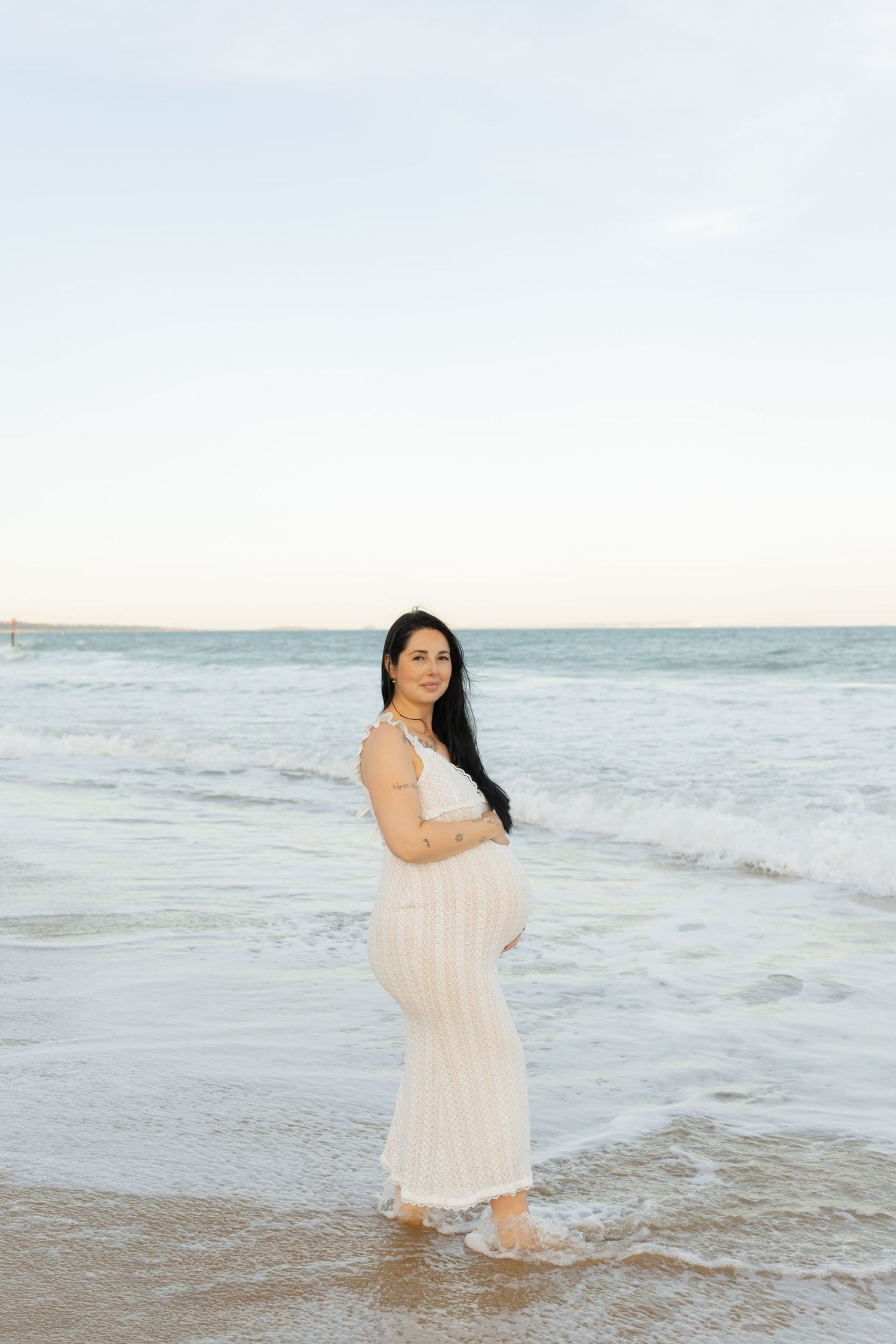Pregnant woman in white dress standing on beach shore with ocean waves, maternity photo session at sunset.