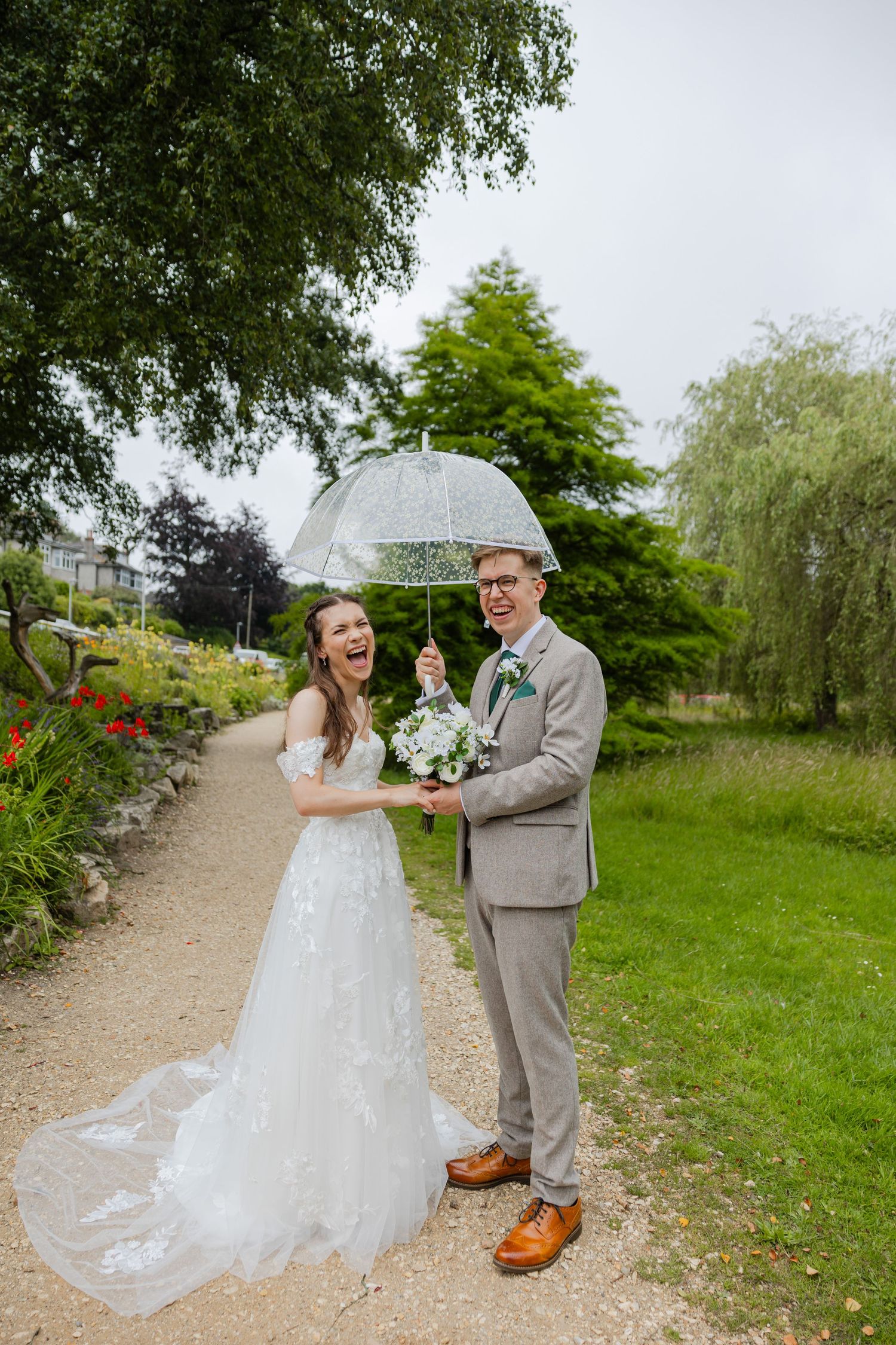 A couple in wedding attire sharing an umbrella on a garden path surrounded by greenery.