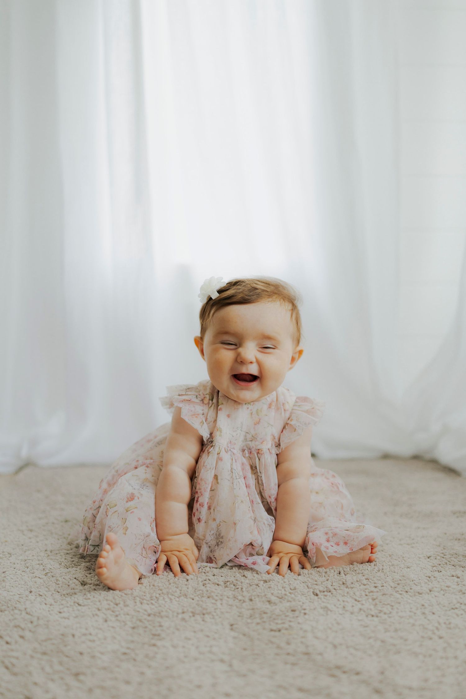 Baby in pink dress laughs while sitting on carpet.