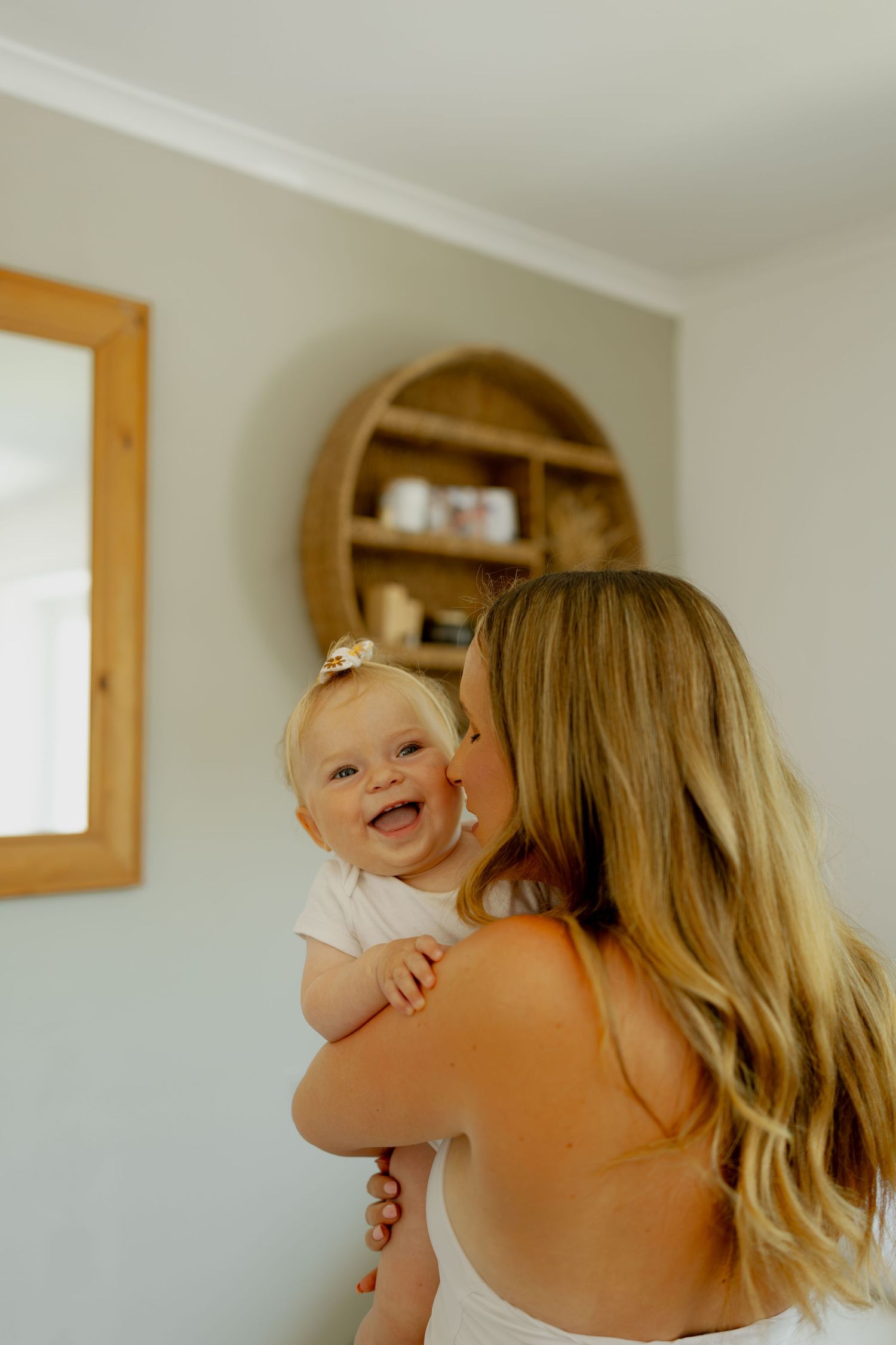 A smiling baby being kissed on the cheek by a blonde mother in a bright, cozy room with wooden decor.