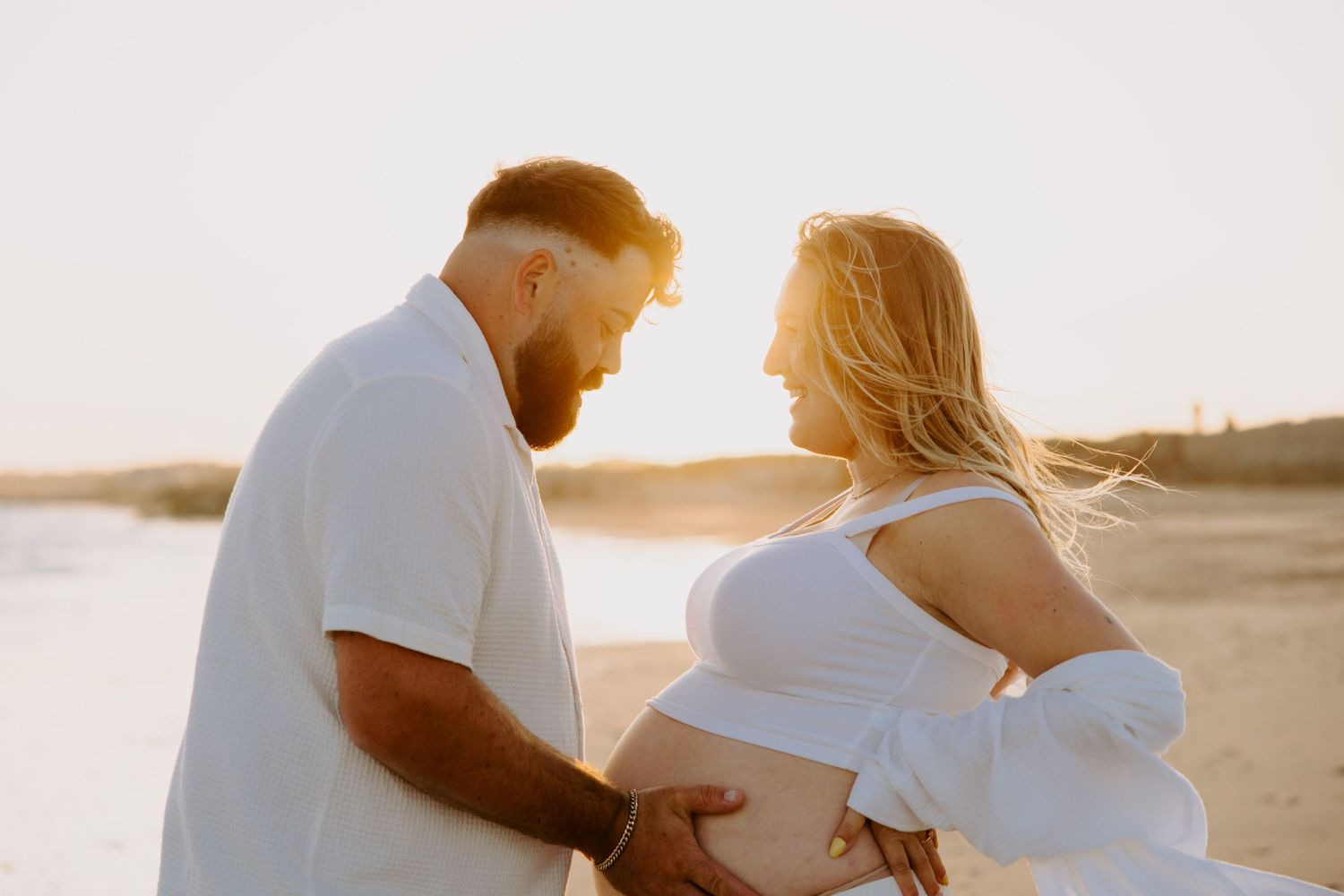 Tender beach maternity photo with couple smiling in golden sunlight.