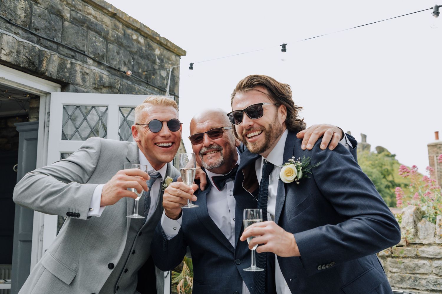 Three men in formal attire toast with champagne glasses at outdoor wedding celebration.