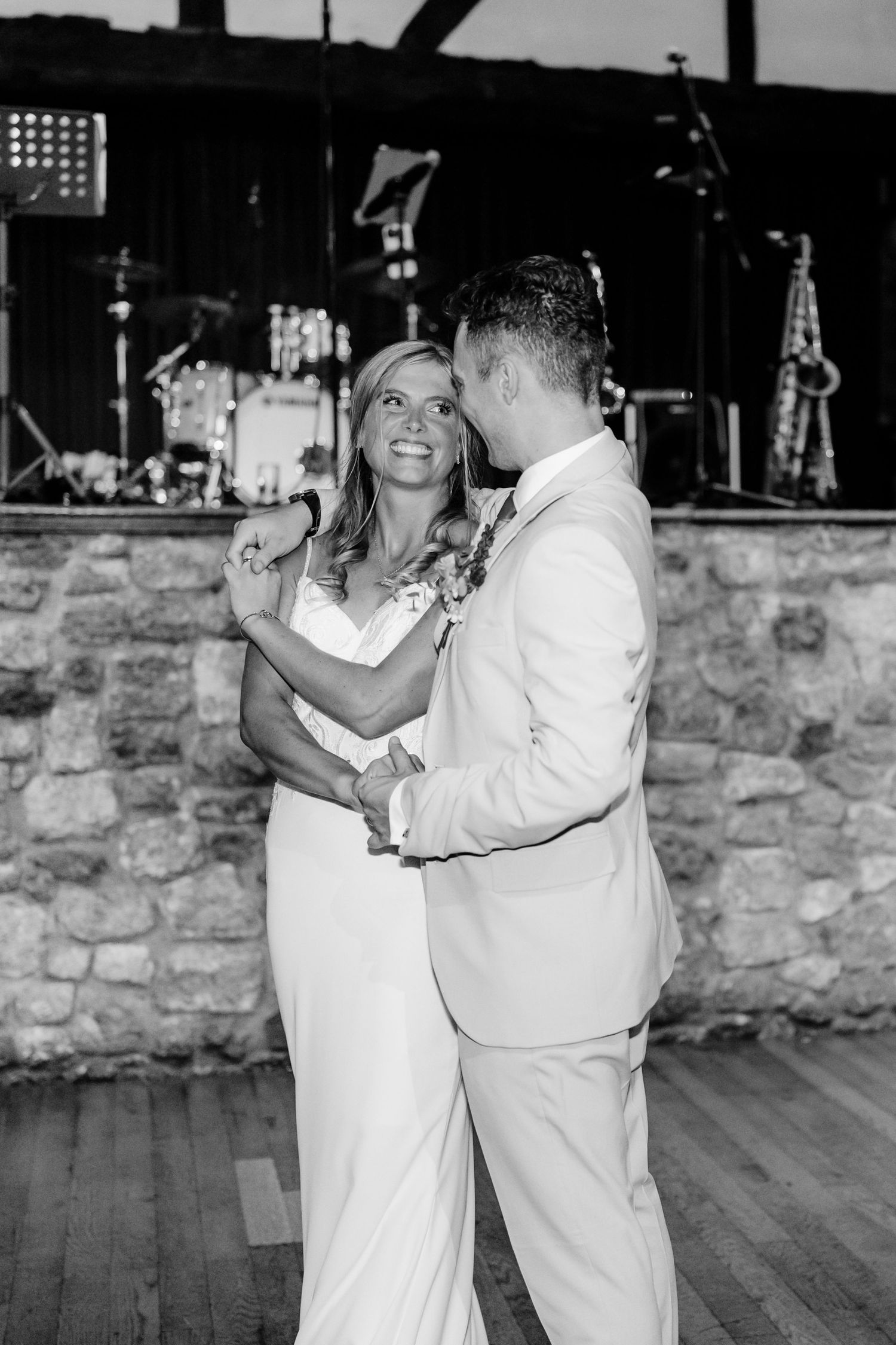 A couple shares their first dance at their wedding reception on a wooden floor against a stone wall backdrop.