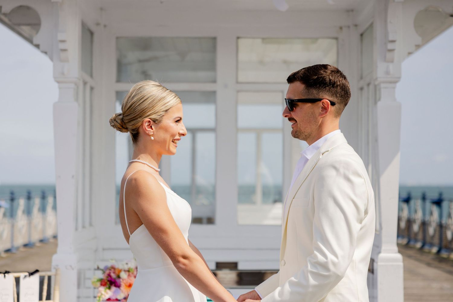 Couple in white wedding attire sharing a joyful moment on oceanfront porch.