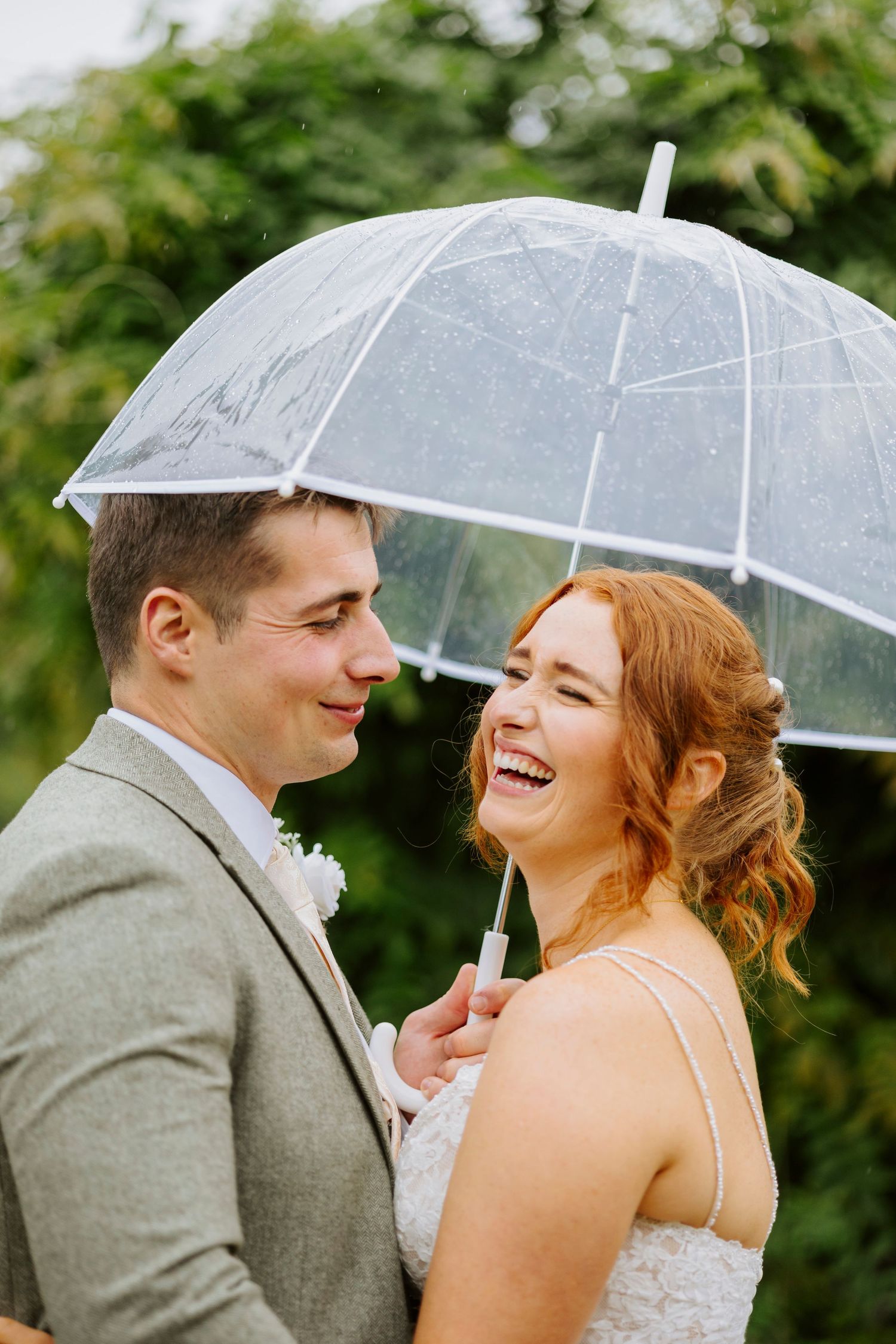 A couple shares a laugh under a clear umbrella during their wedding photoshoot.