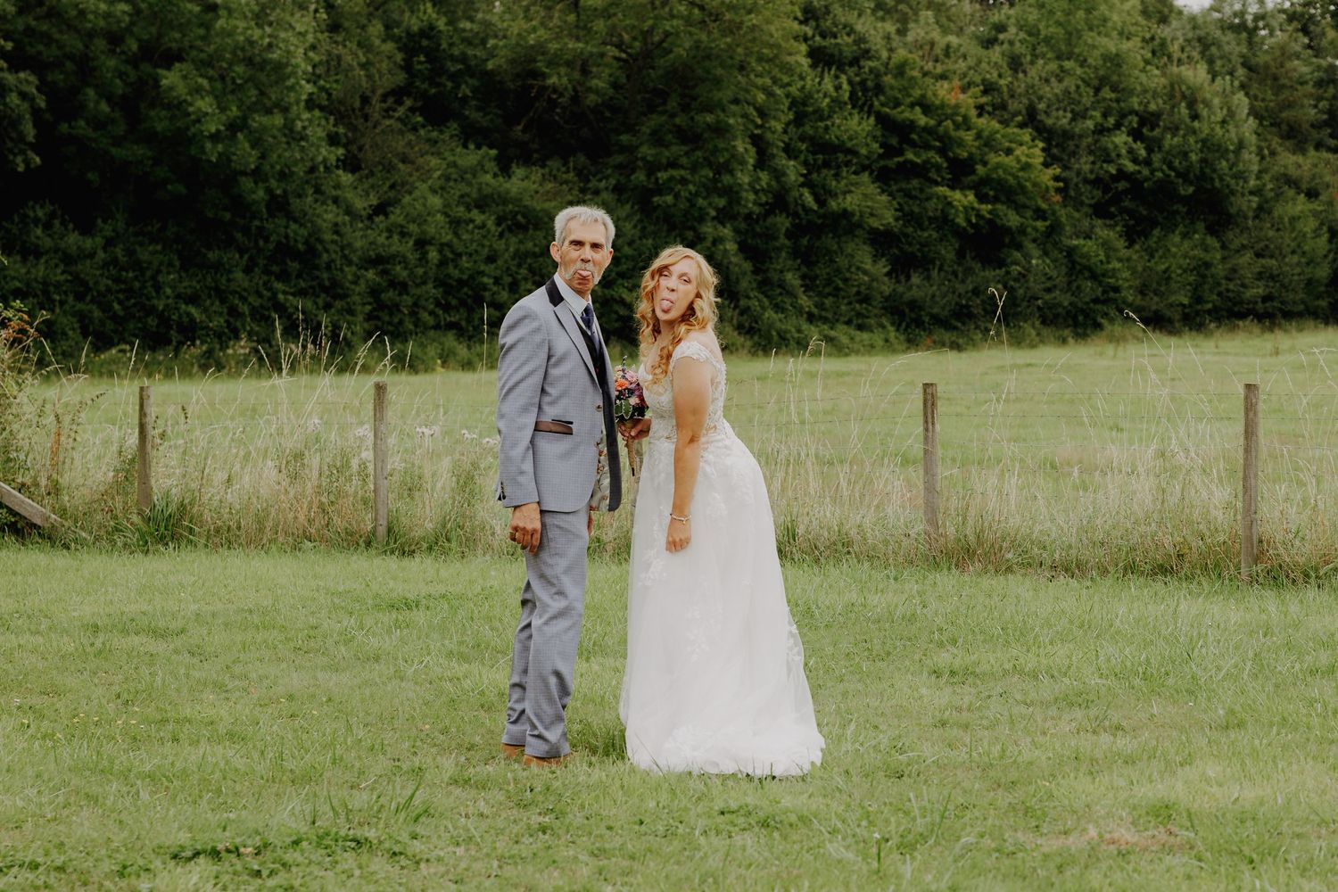 Bride and groom in formal attire pose together in a sunny meadow.