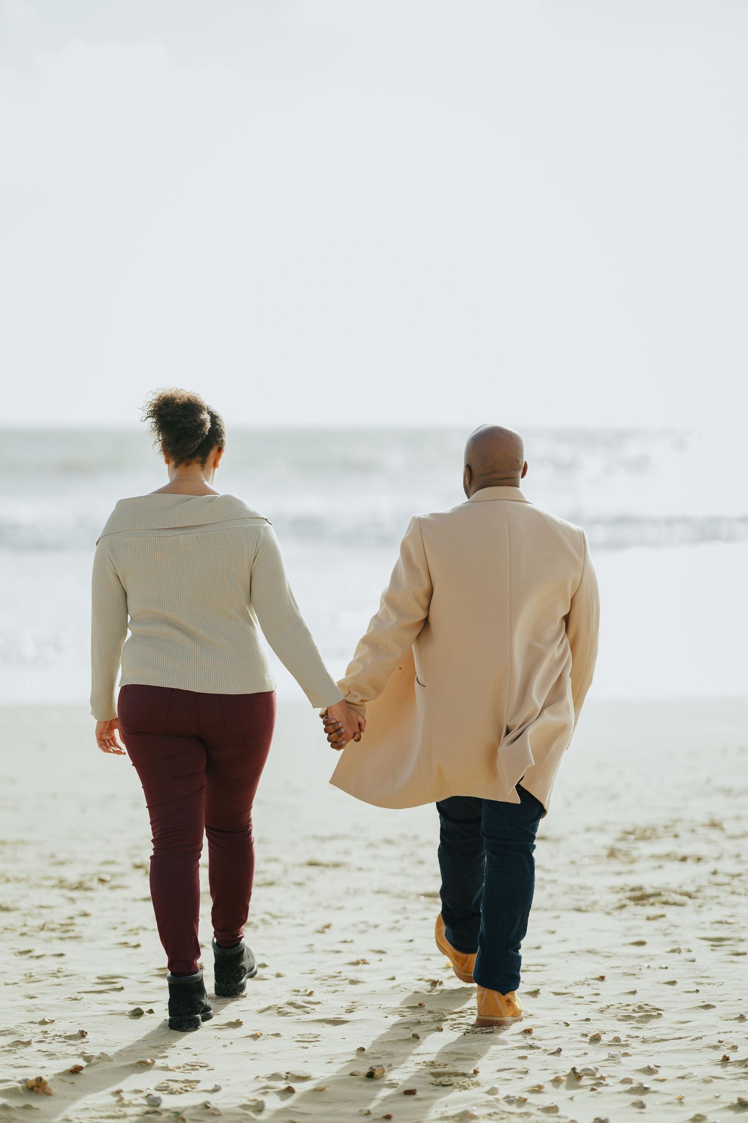 A couple in matching neutral tones walks hand in hand along a misty beach.