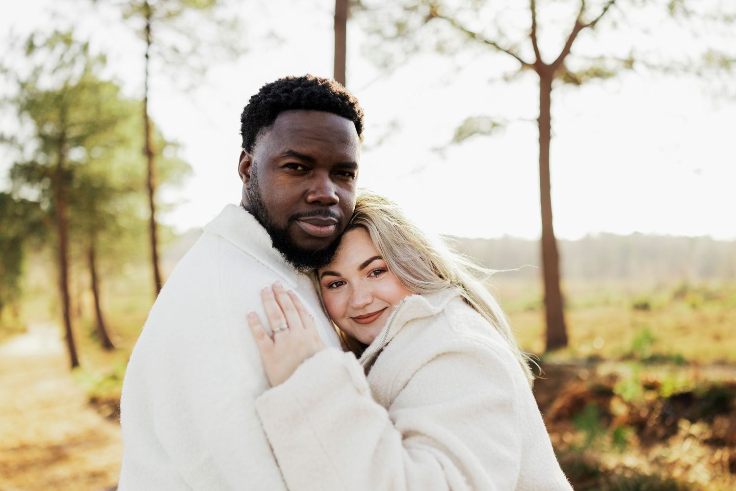 A couple wearing matching cream sweaters embrace outdoors in a scenic pine forest setting during golden hour.