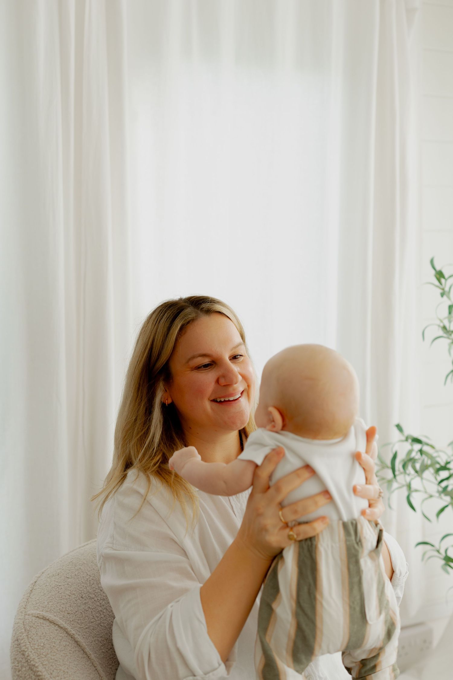 A tender moment between parent and infant in a bright white room with sheer curtains and greenery.
