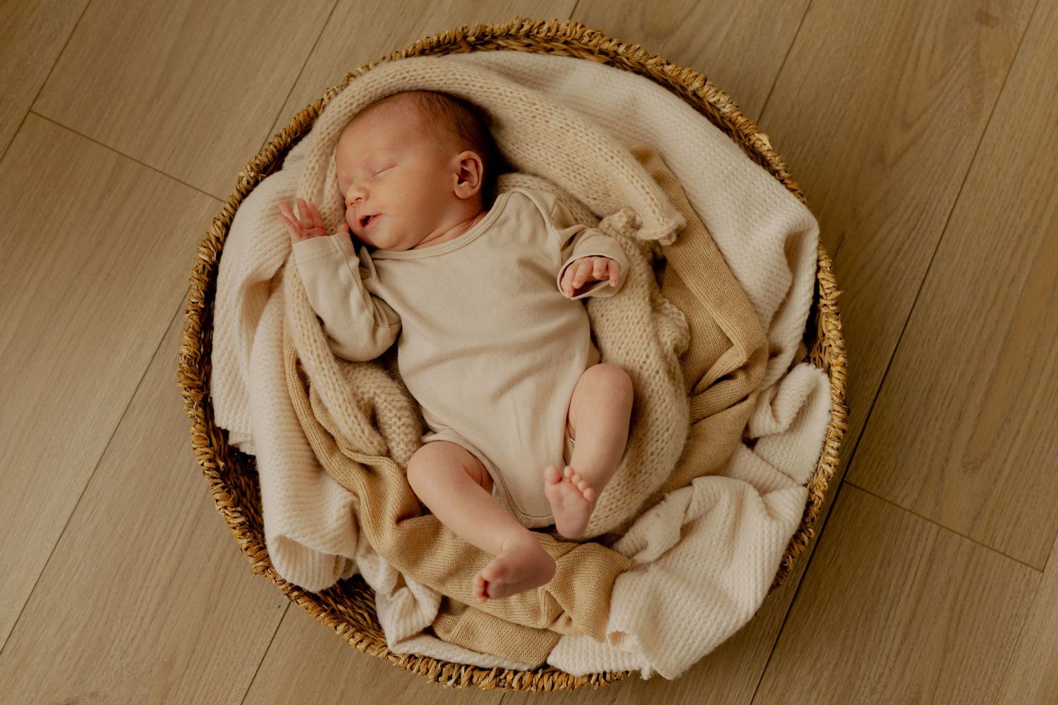 A sleeping newborn baby lies curled up on a cream-colored blanket in a round woven basket on wooden flooring.