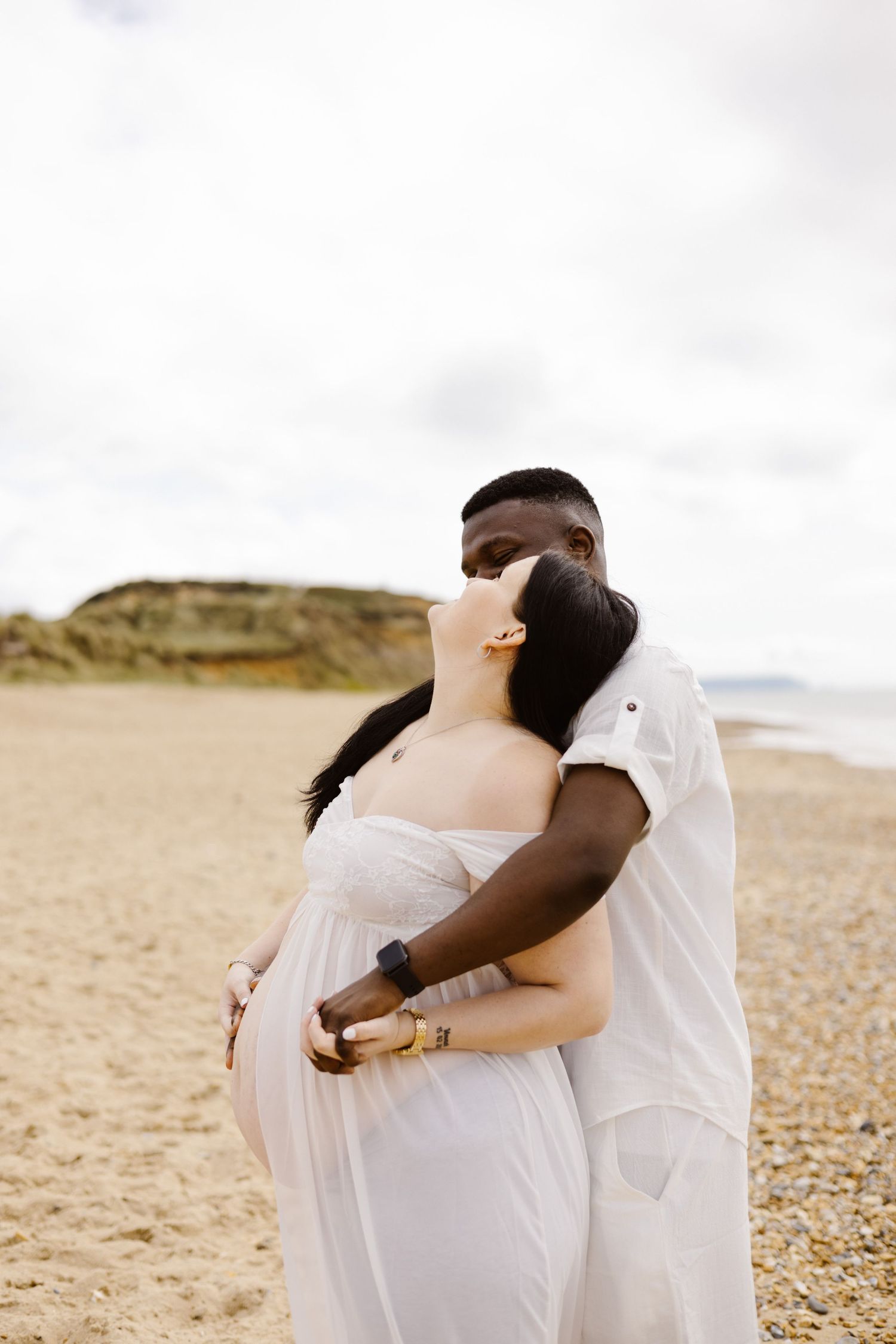A tender moment between expecting parents embracing on a cloudy beach.