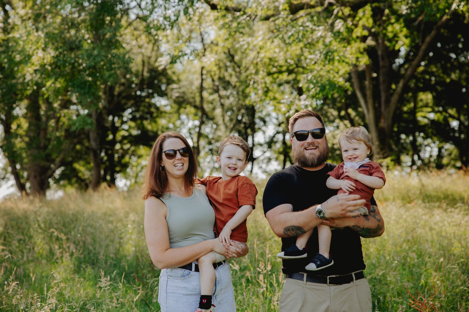 Family portrait of parents with two children in a sunny meadow surrounded by trees.