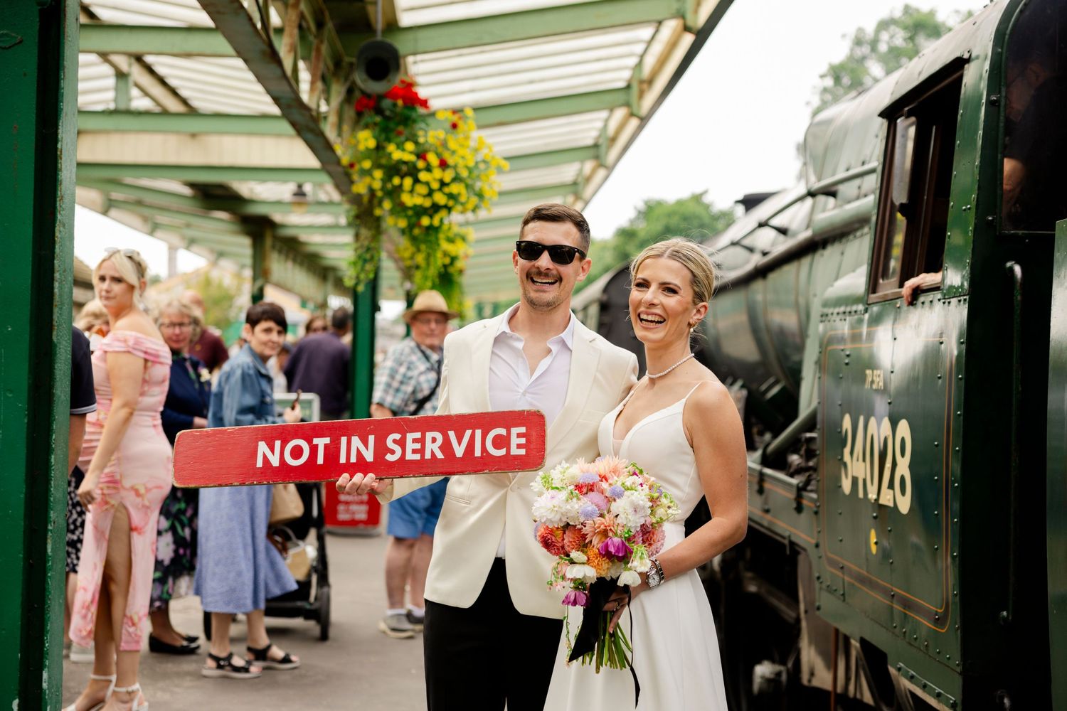 A couple in wedding attire stands beside a vintage train with a 'Not in Service' sign.