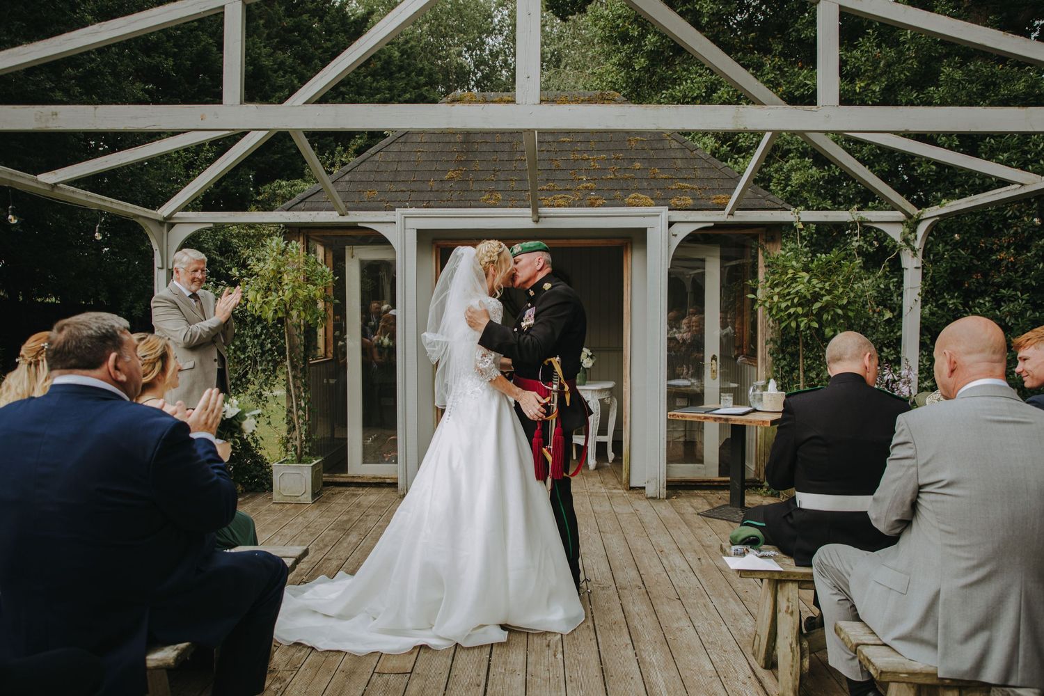 Couple kisses during outdoor wedding ceremony under white gazebo with seated guests watching.