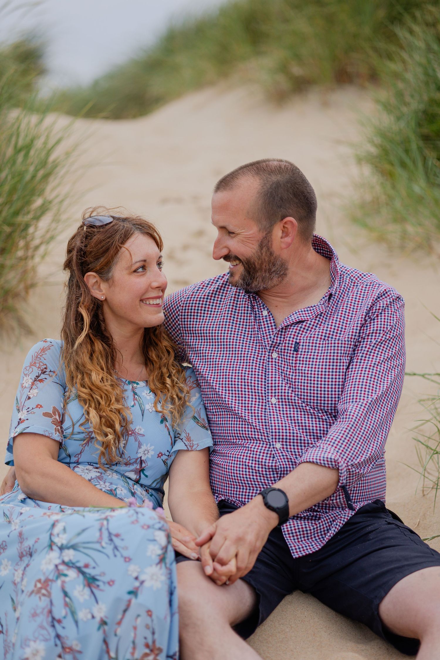 A couple sits together on sandy dunes surrounded by beach grass.