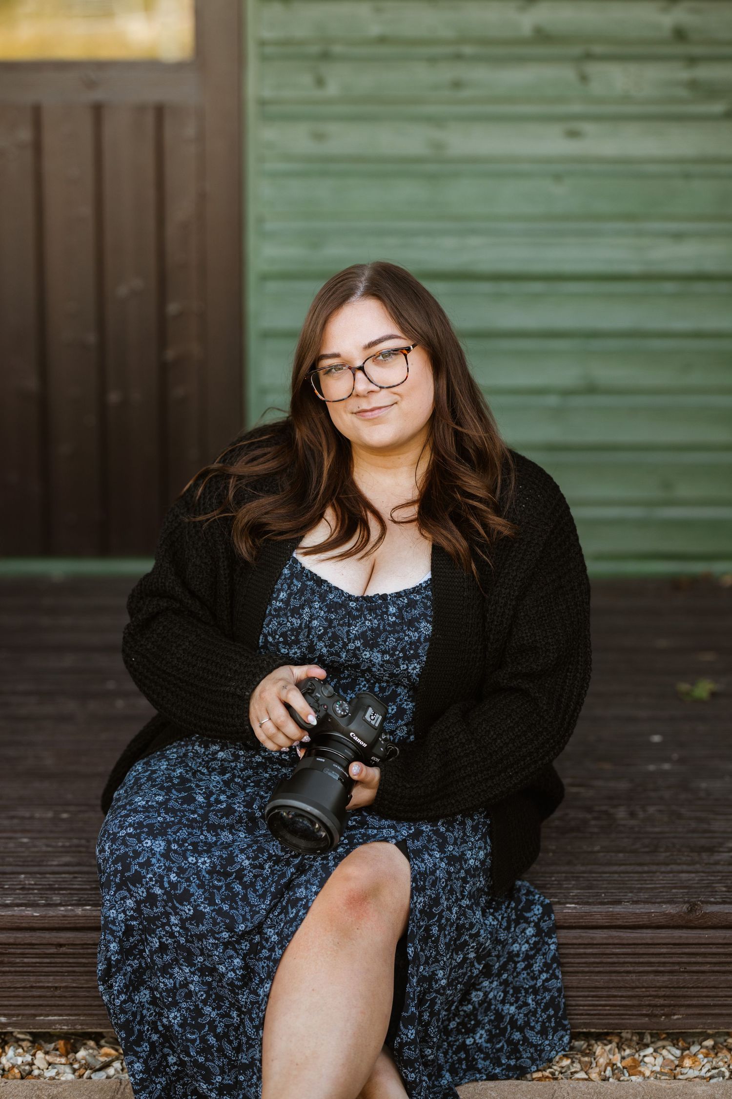 A photographer in glasses and a floral dress holds a camera while sitting in front of a green wall.