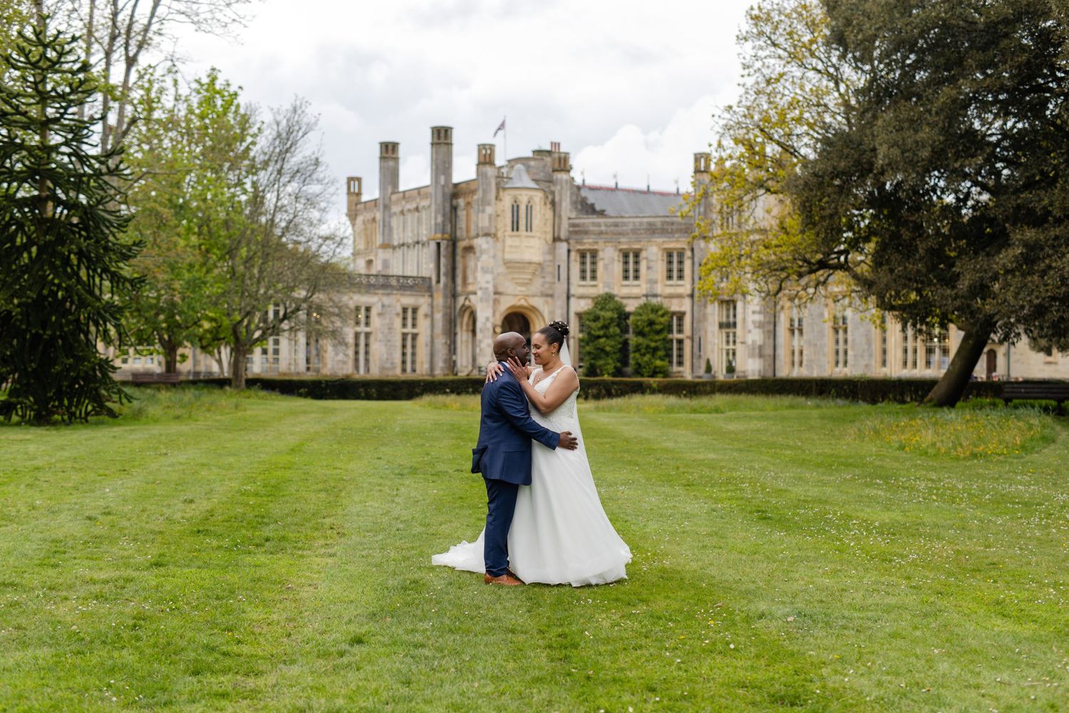 A couple embraces on the lawn in front of a grand historic mansion on their wedding day.