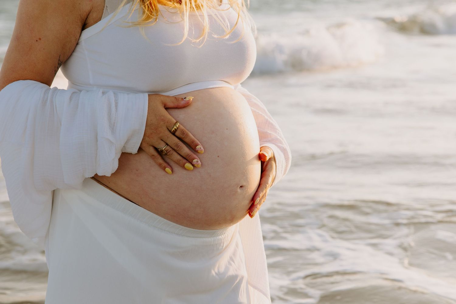 Pregnant woman in white dress cradling her belly on beach with waves.