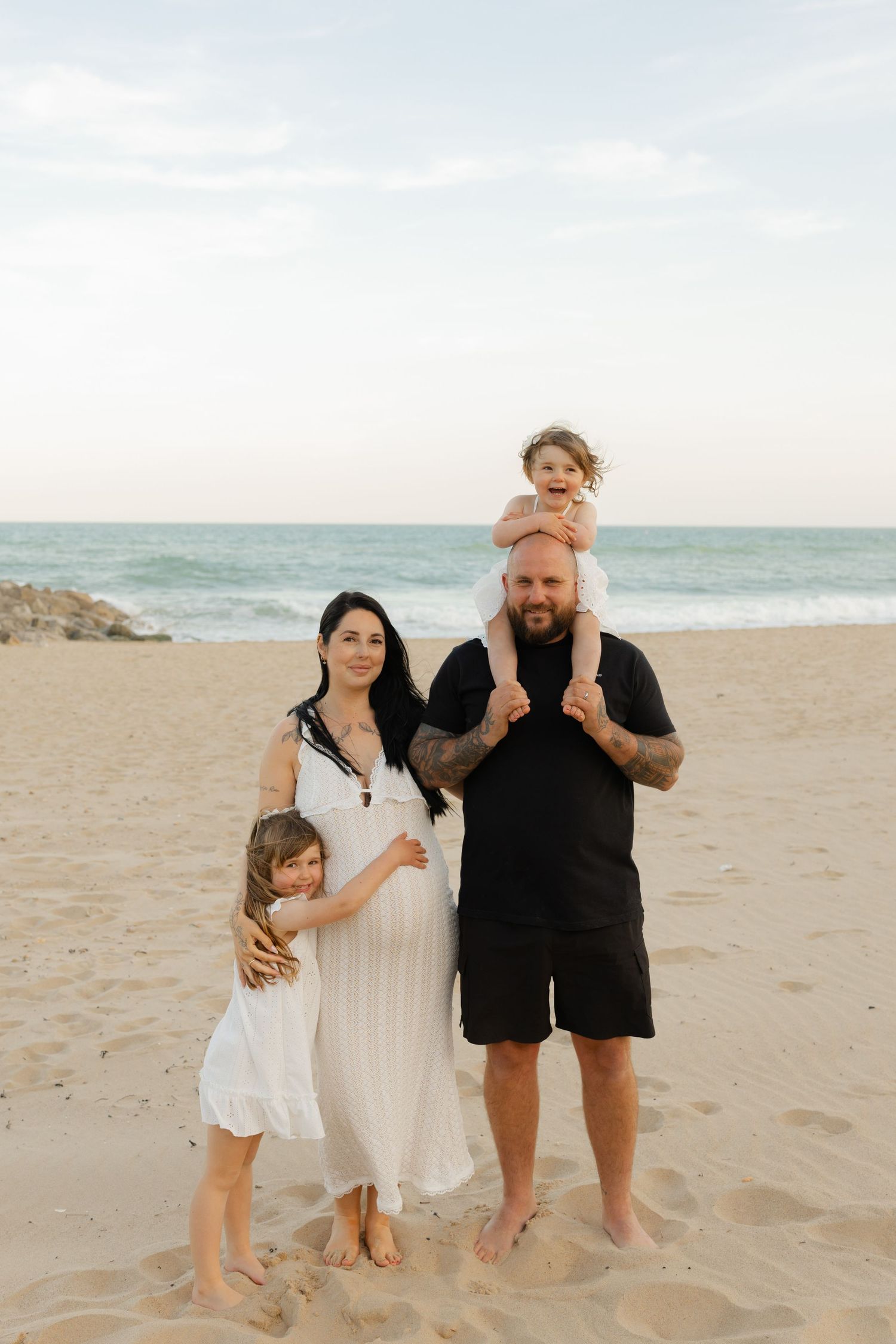 Family of four poses on sandy beach at sunset, pregnant mother in white dress, tattooed father with toddler on shoulders.