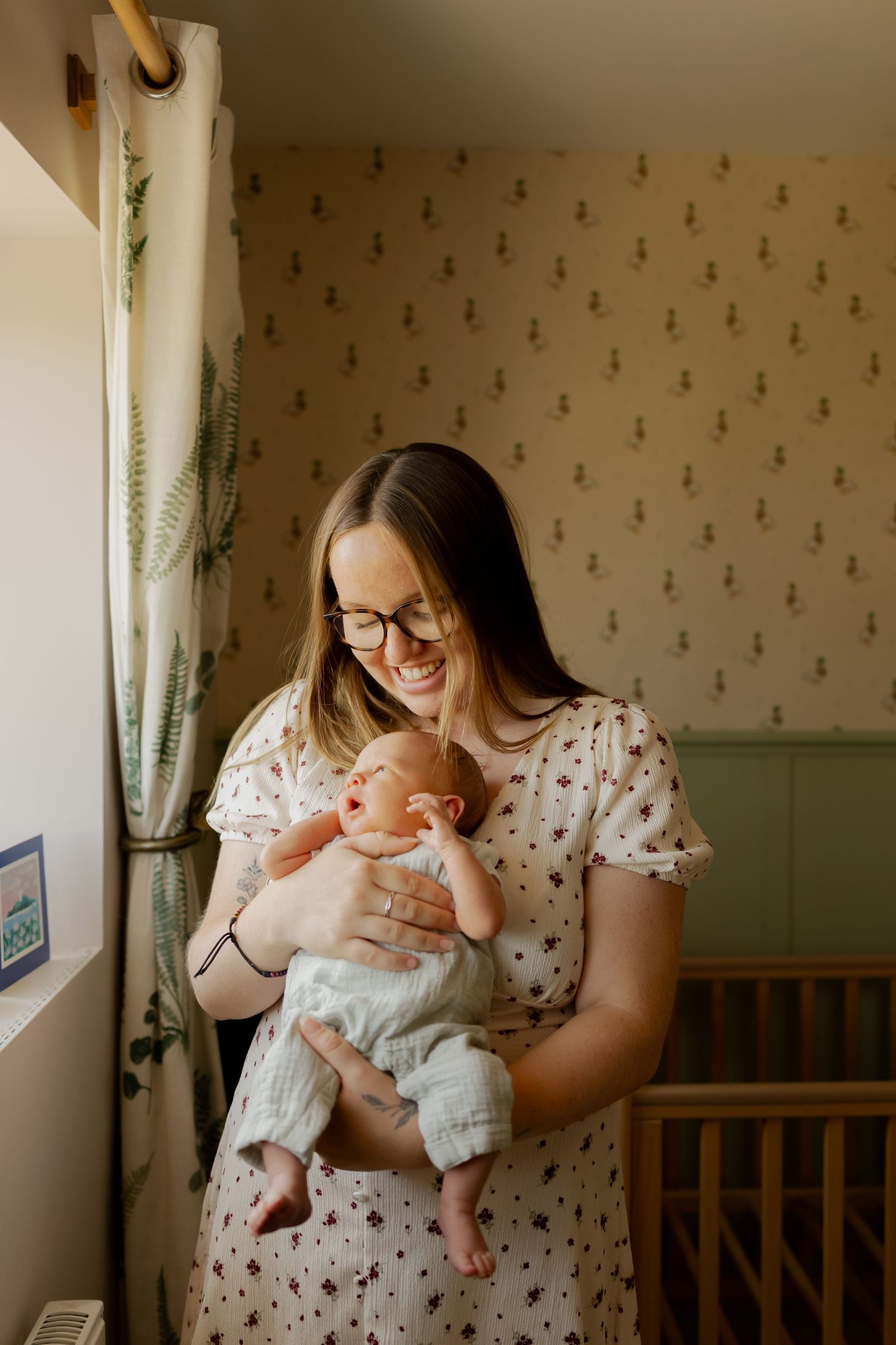 Smiling mother in floral dress holding newborn baby in a nursery room with patterned wallpaper.