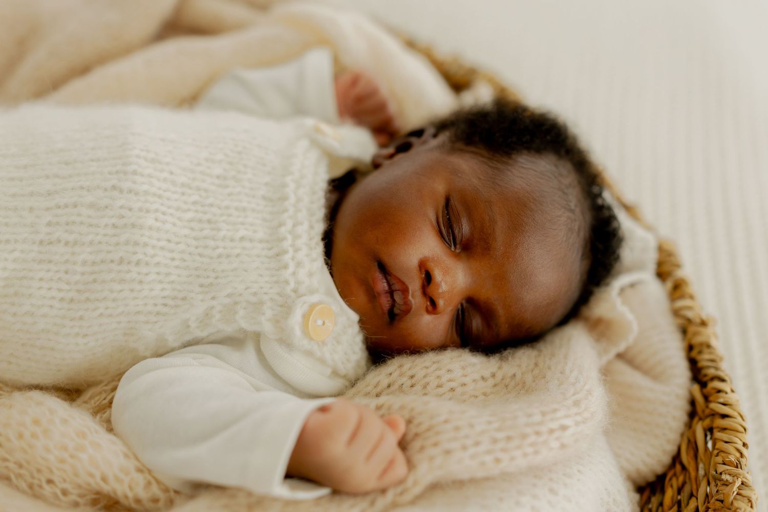 Peaceful newborn baby sleeping in a wicker basket, wrapped in a cream knit sweater and soft blanket.