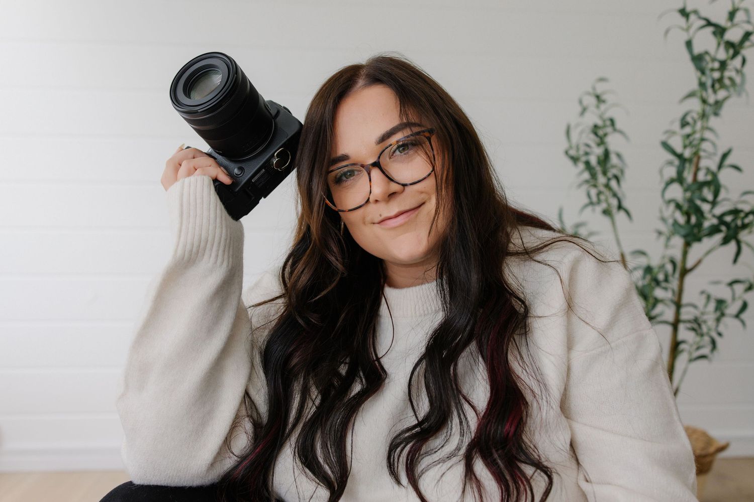 A person with long dark hair and glasses poses playfully in a bright white room with green plants.