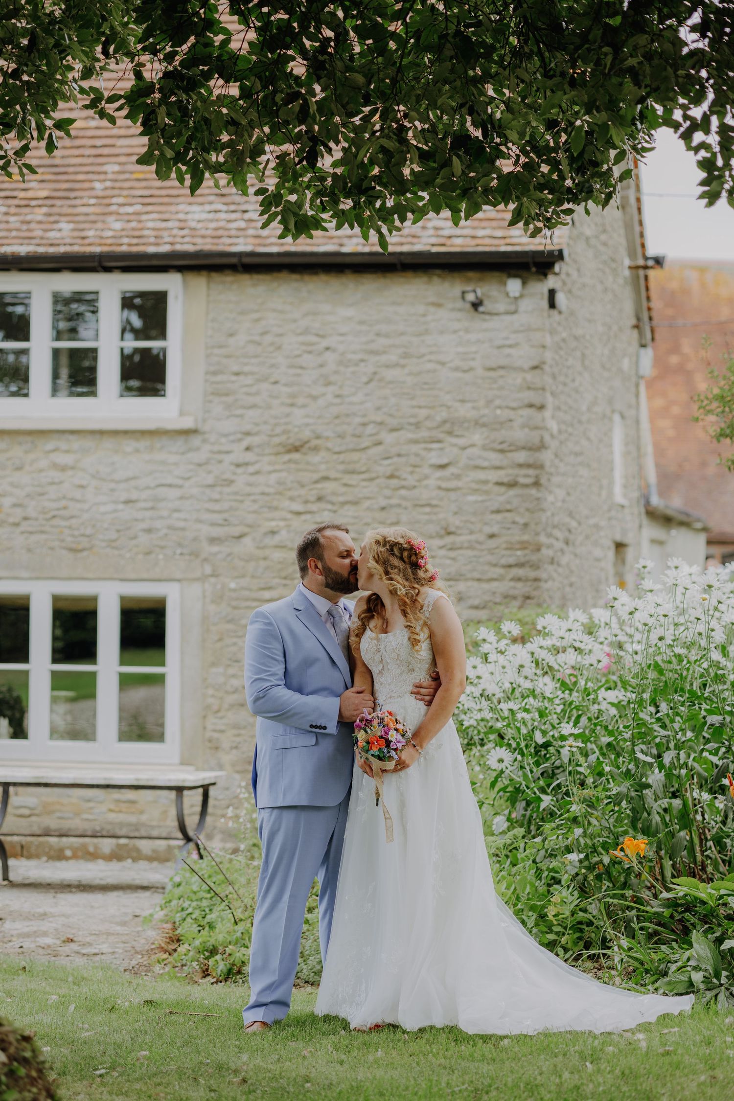 Newlyweds in light blue suit and white dress pose in front of stone house with garden.