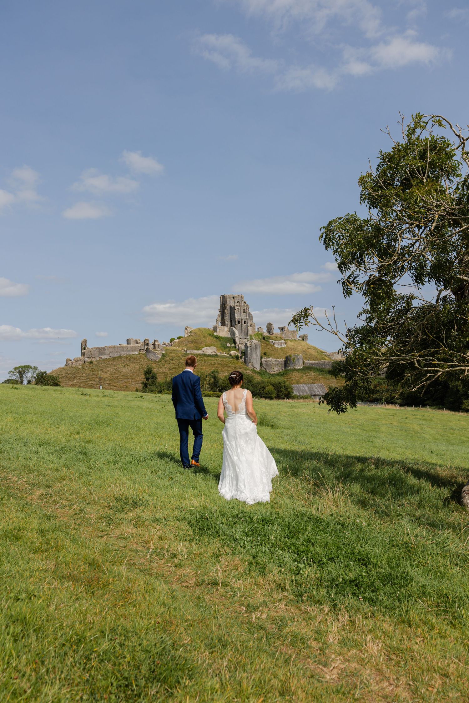A wedding couple walks toward an ancient castle ruin atop a grassy hill under blue skies.