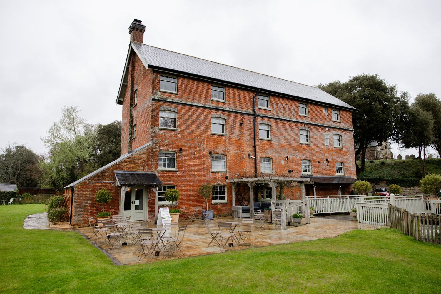 A historic red brick mill building with multiple stories sits on a grassy lawn surrounded by trees.