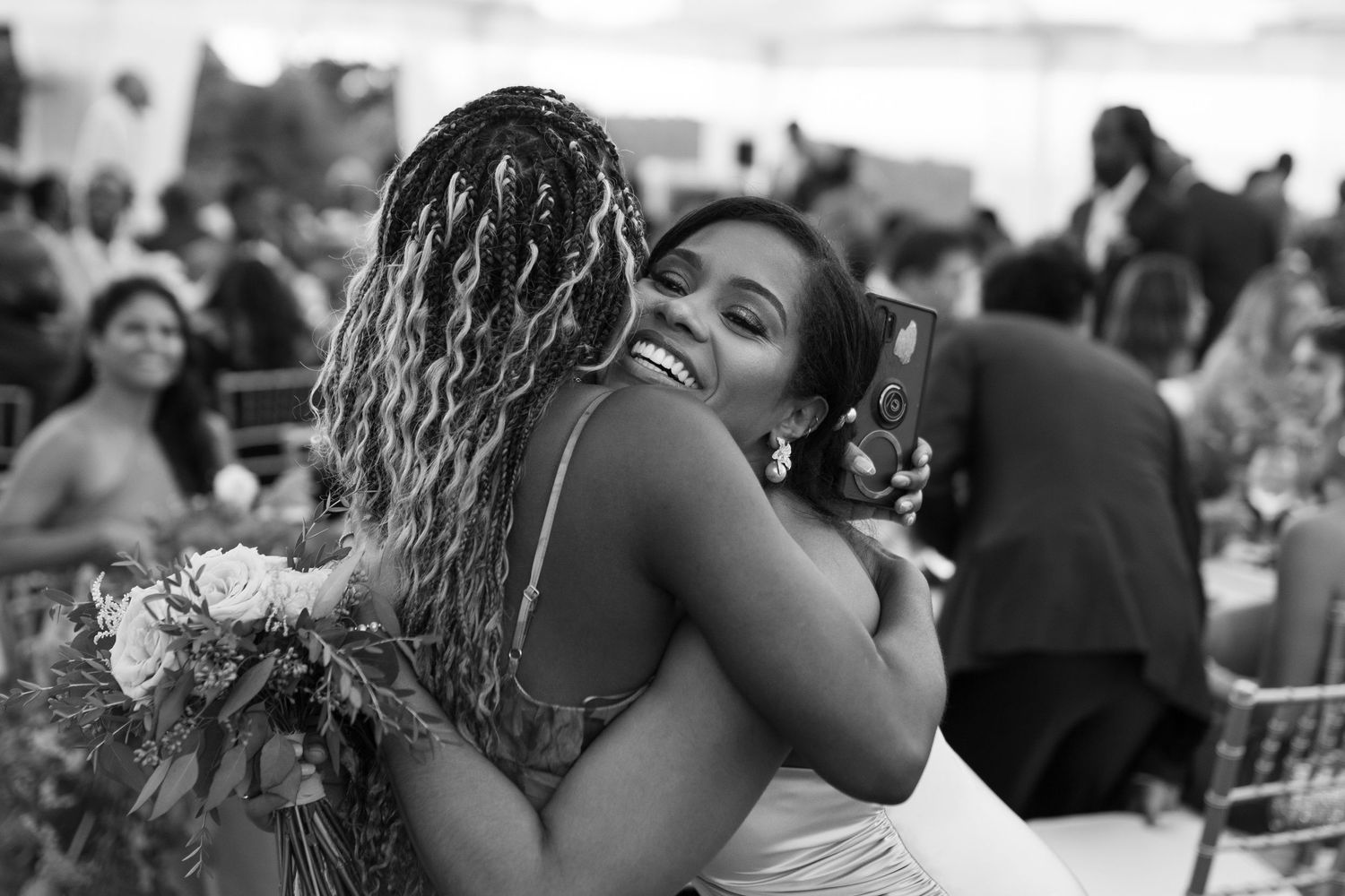 Two people sharing an emotional embrace at an outdoor celebration while holding a floral bouquet in black and white.