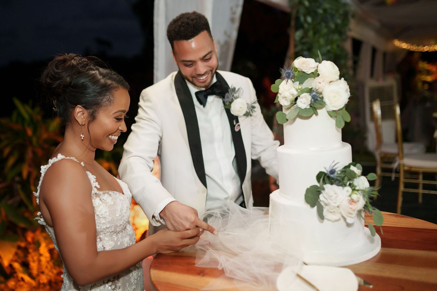 Newlyweds cut their elegant white tiered wedding cake adorned with white roses and greenery at reception.
