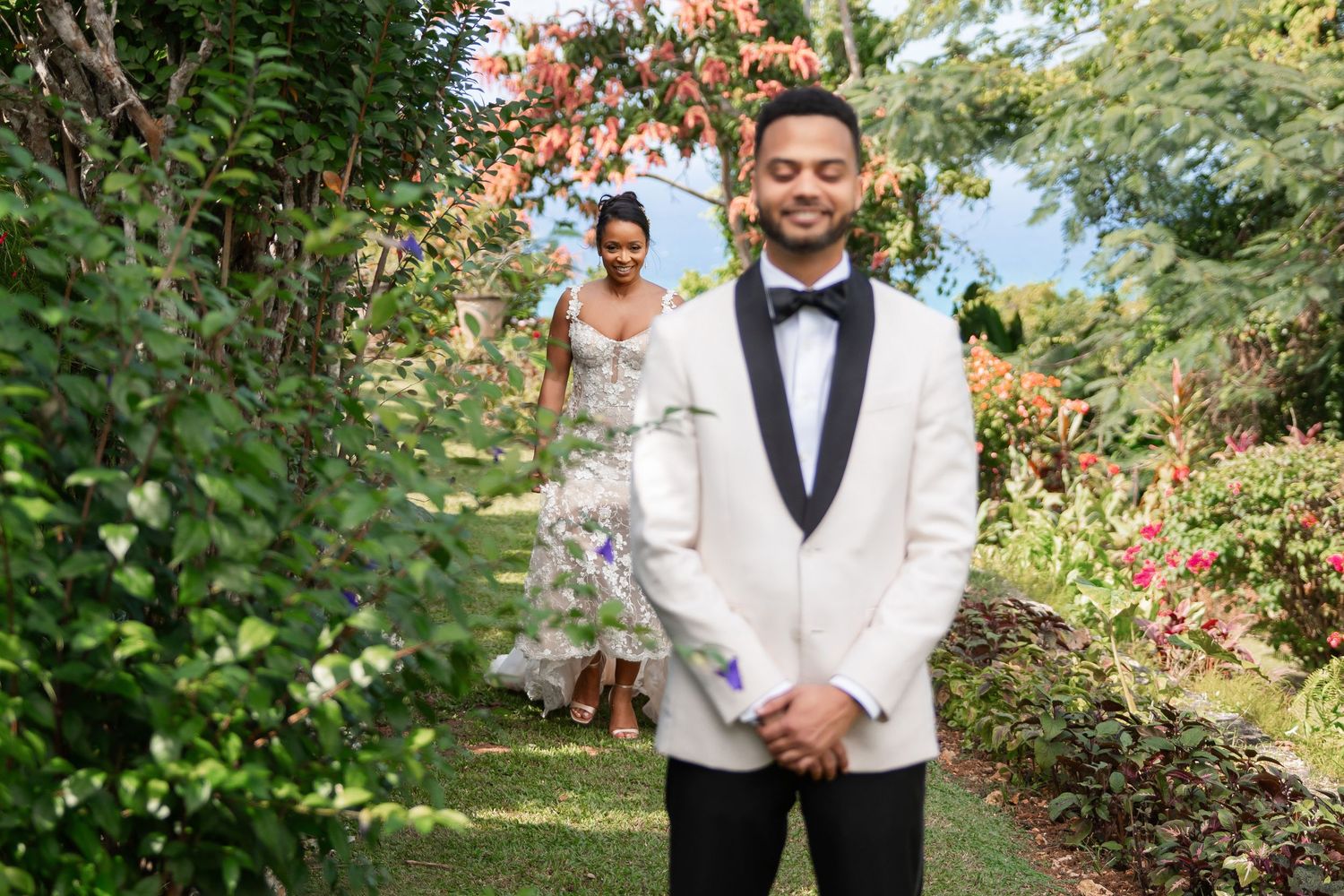 A groom in a classic white dinner jacket and black bow tie poses in a lush garden setting before the ceremony.