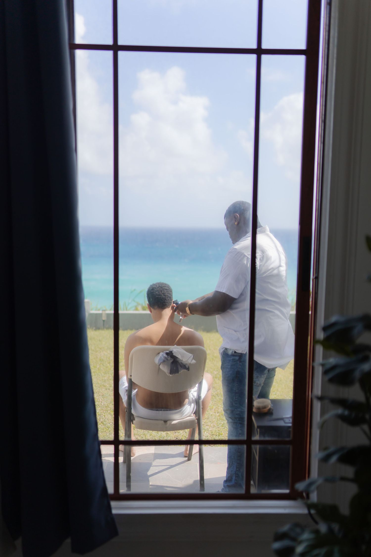 Two people sit on chairs looking out through large windows at a tropical ocean view with bright turquoise water.