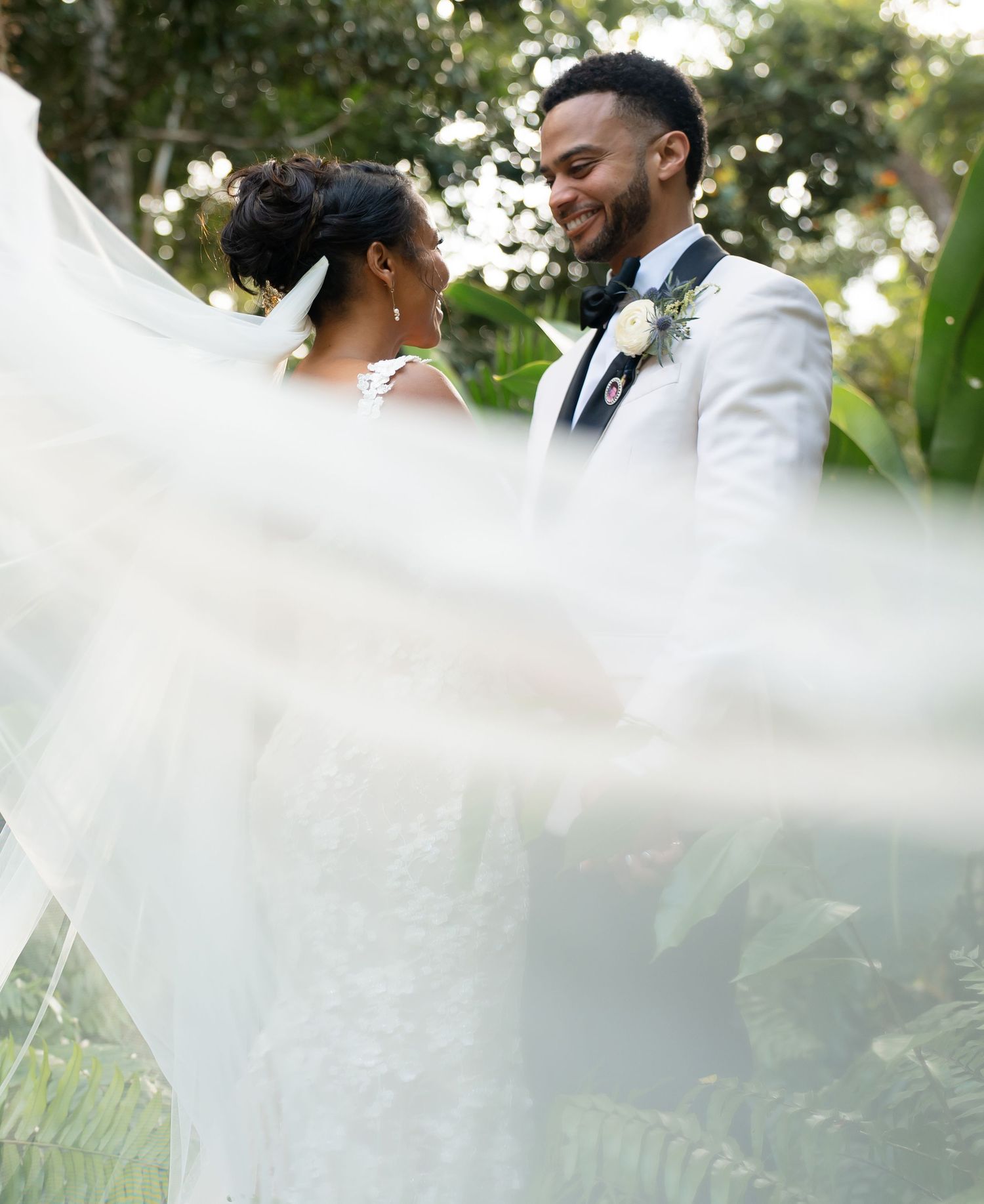 A joyful newlywed couple share a tender moment in a lush garden setting with flowing white veil in the foreground.