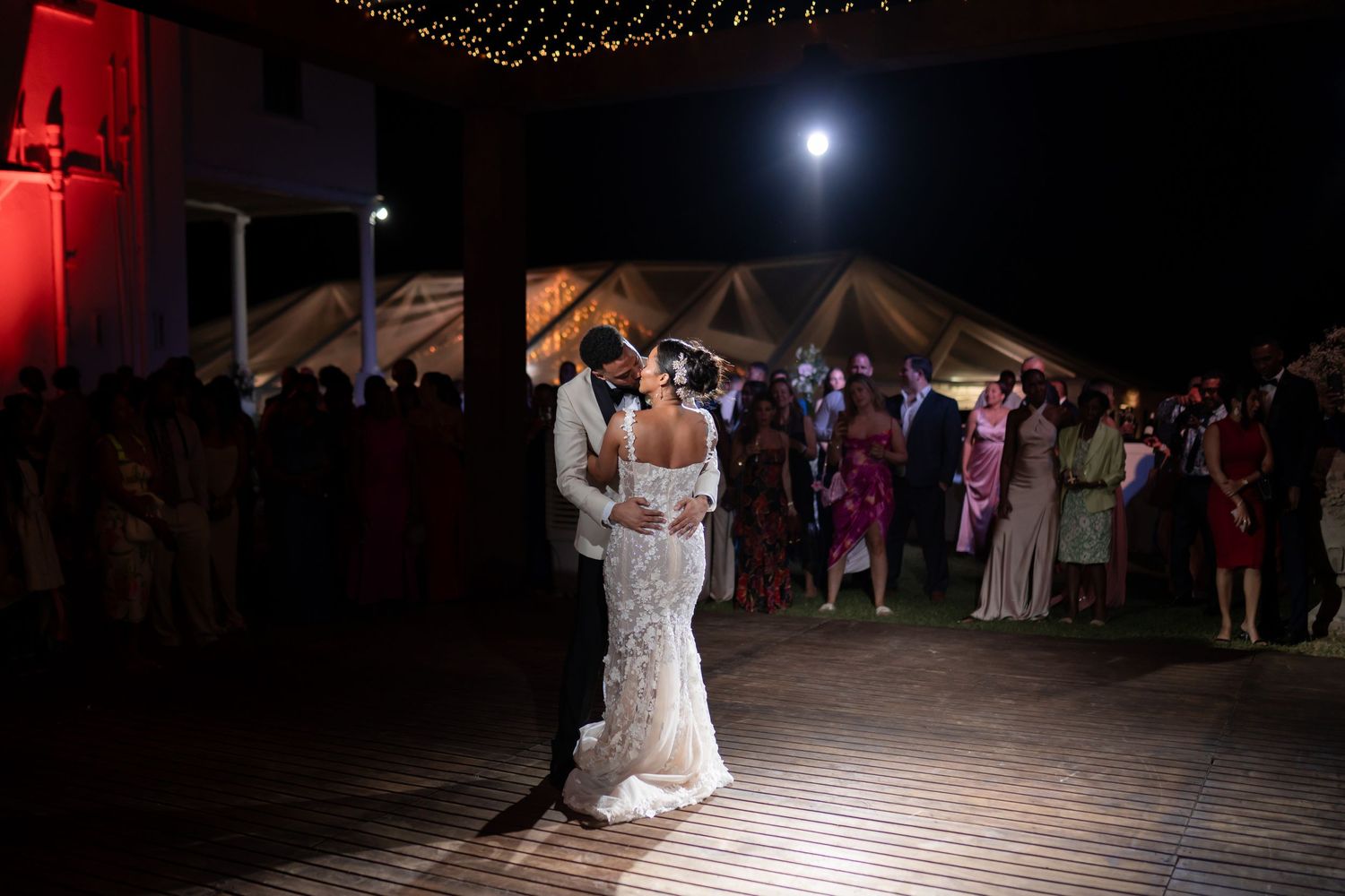 A couple shares their first dance at a wedding reception under a tent with guests watching and a full moon in the background.