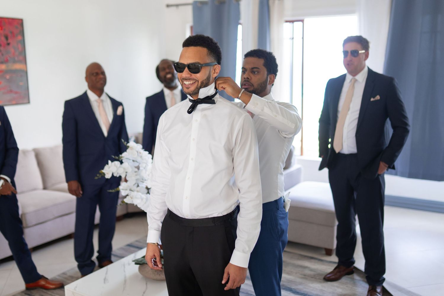 A groom in a crisp white shirt and bowtie adjusts his outfit while getting ready in a bright modern room.