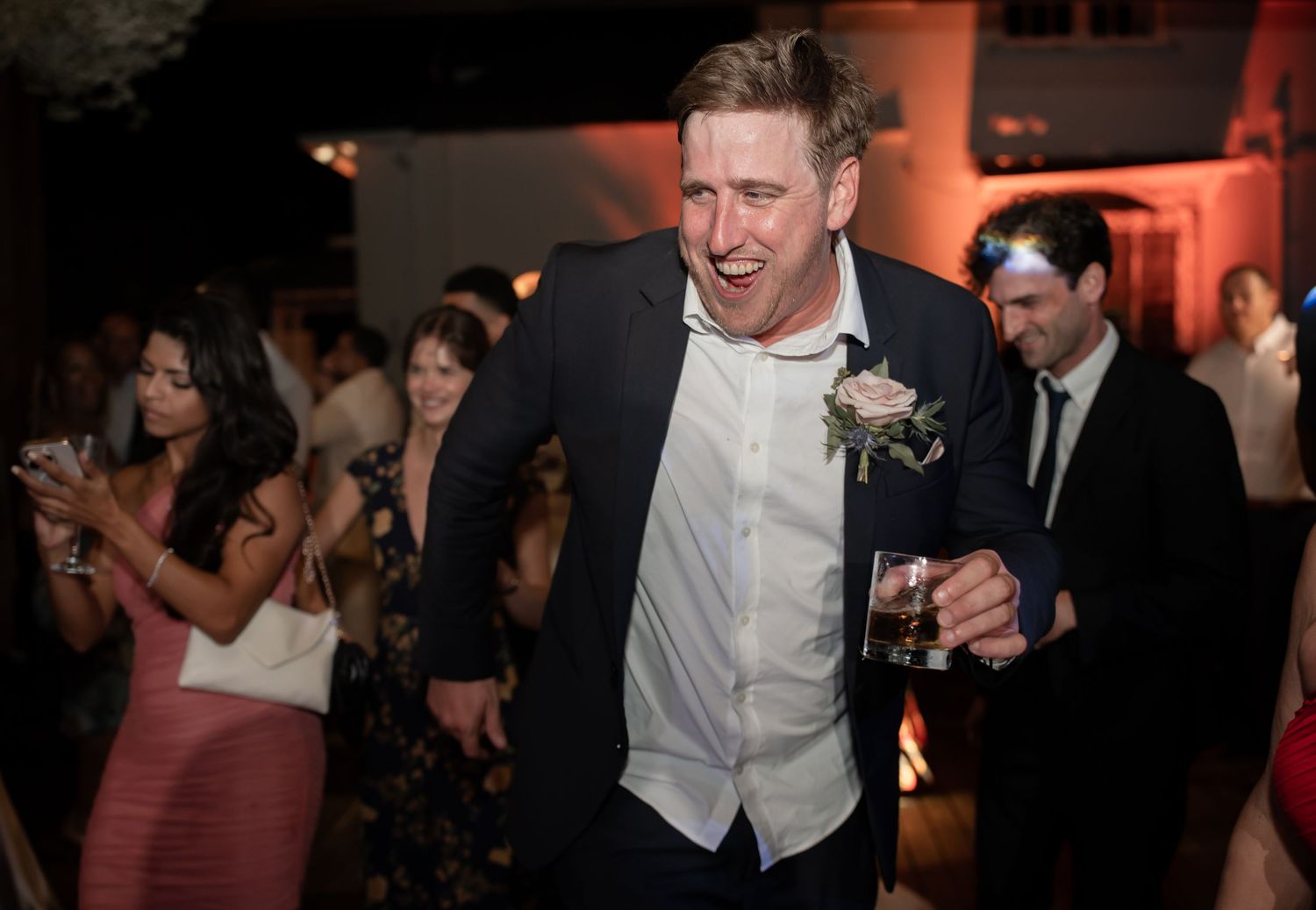 Wedding guests dance and celebrate together on a dimly lit dance floor with warm red lighting at a reception venue.